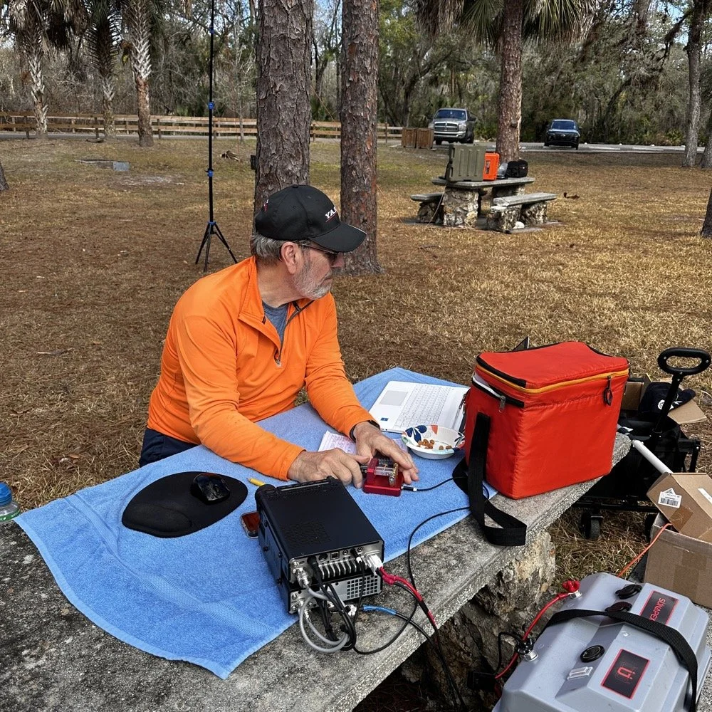Amateur radio operator in orange shirt operating Yaesu transceiver at picnic table during multi-operator POTA activation