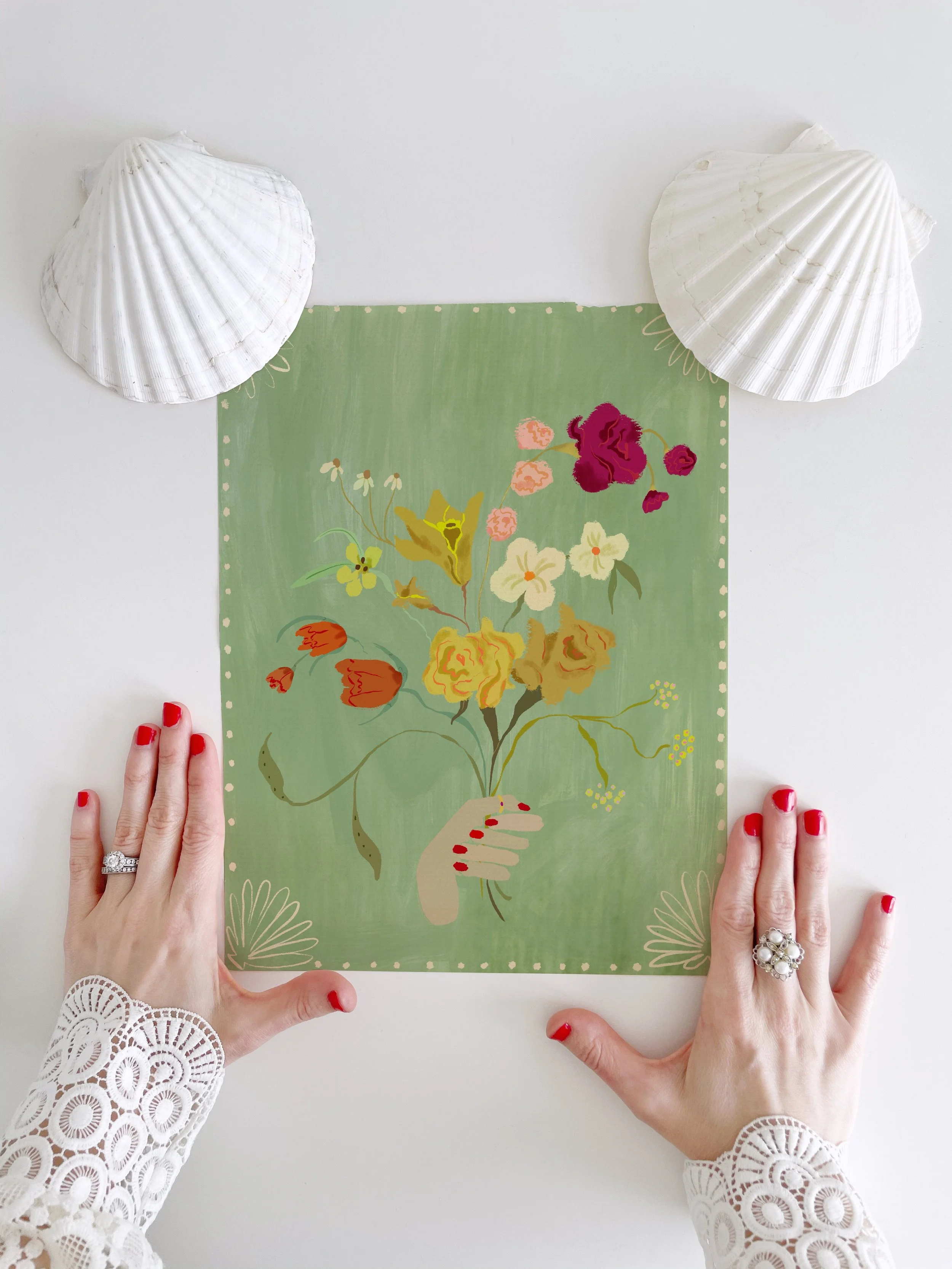 Painterly floral illustration held by a woman’s hand – watercolor wildflower bouquet with lyrical stems and petals, sitting on a white surface, under a ladies manicured hands and two scallop shells