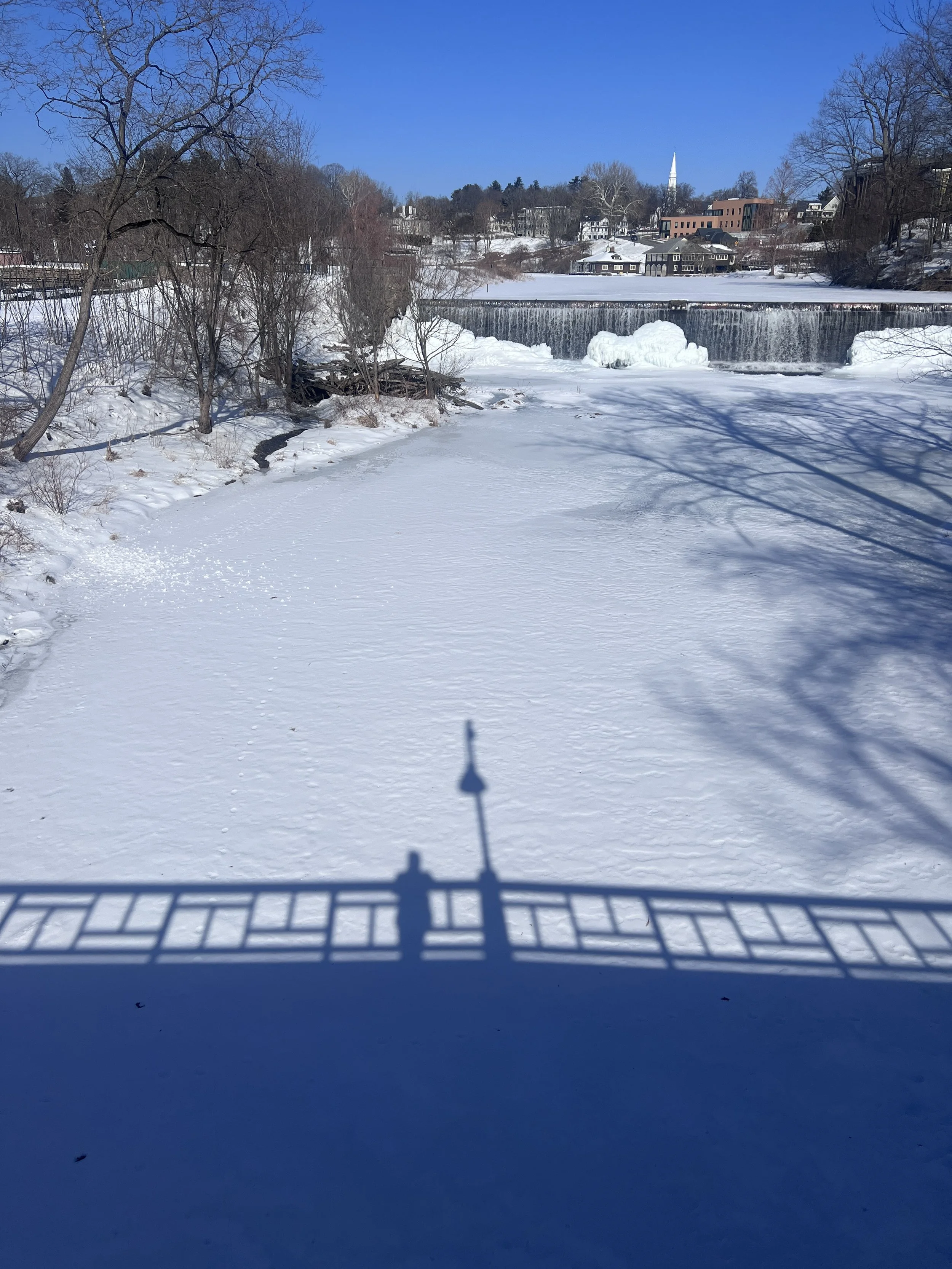View from a snow-covered balcony looking out over a river with a waterfall and houses in the background on a clear, sunny winter day.