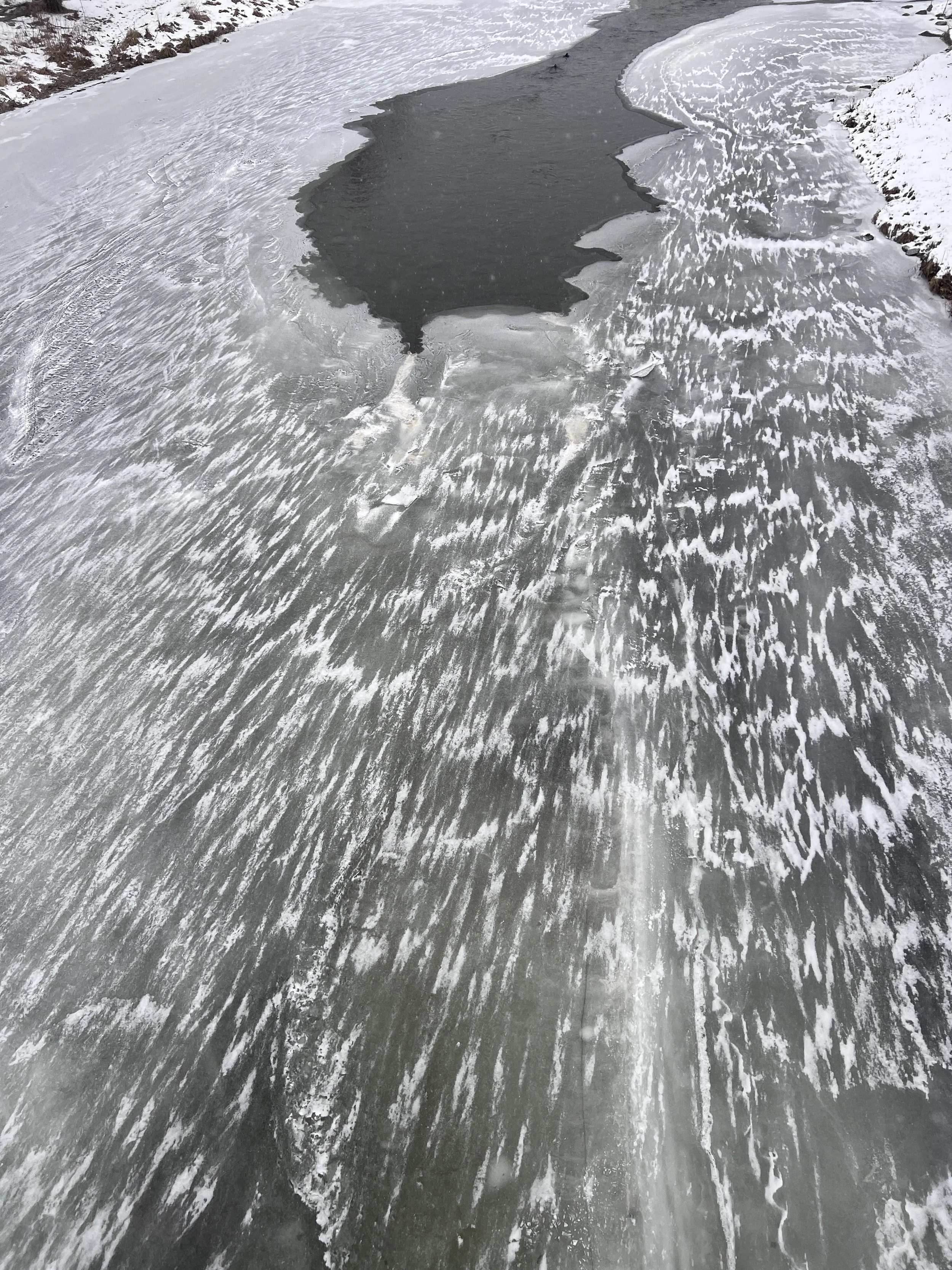 Frozen river with partially frozen and flowing water, surrounded by snow-covered banks and rocky terrain.