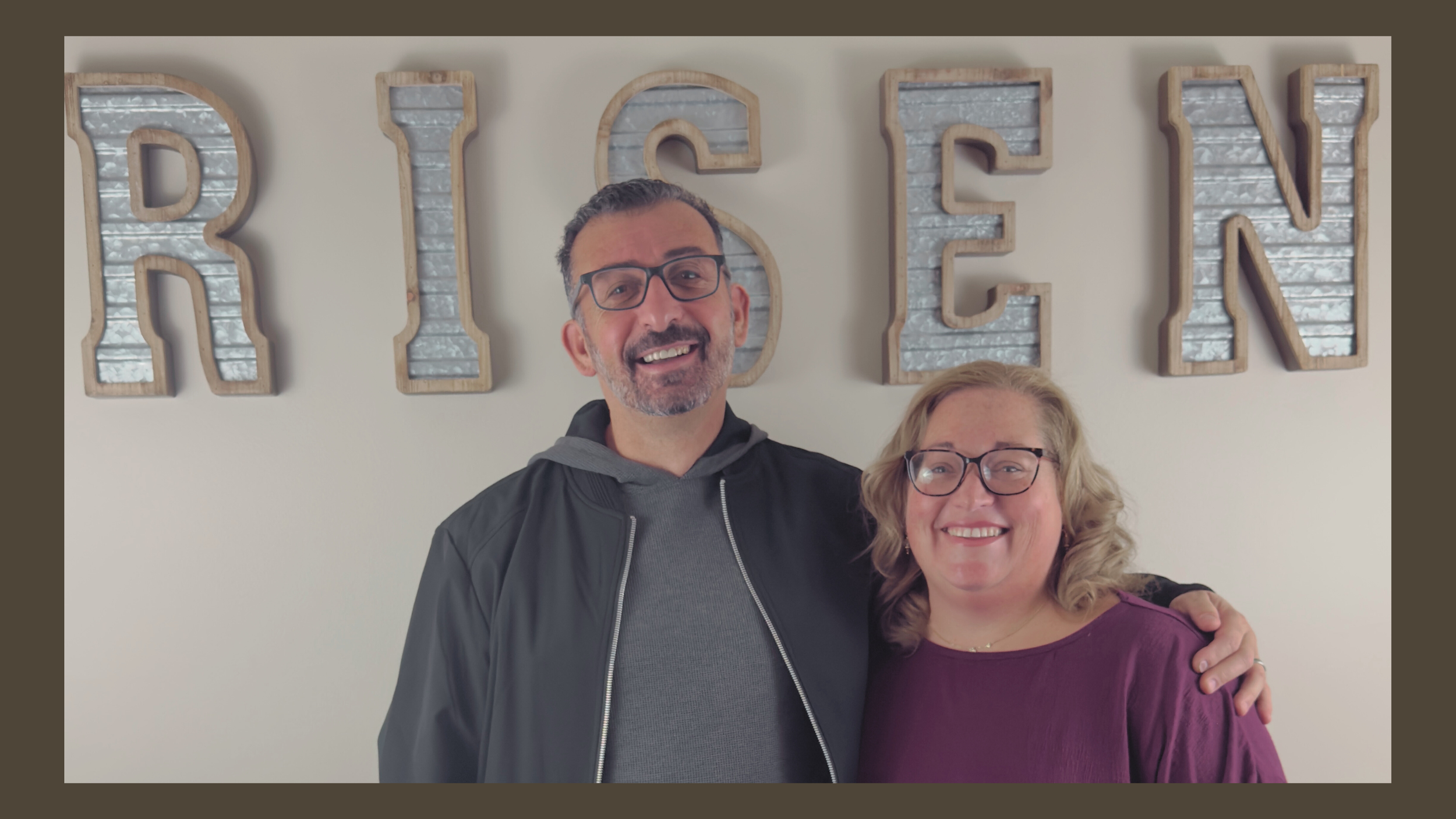 Two people, a man and a woman, smiling and standing close together in front of a wall with large decorative letters spelling "RISON". The man is wearing glasses, a black jacket, and a gray hoodie, while the woman has glasses, curly blonde hair, and is wearing a purple top.