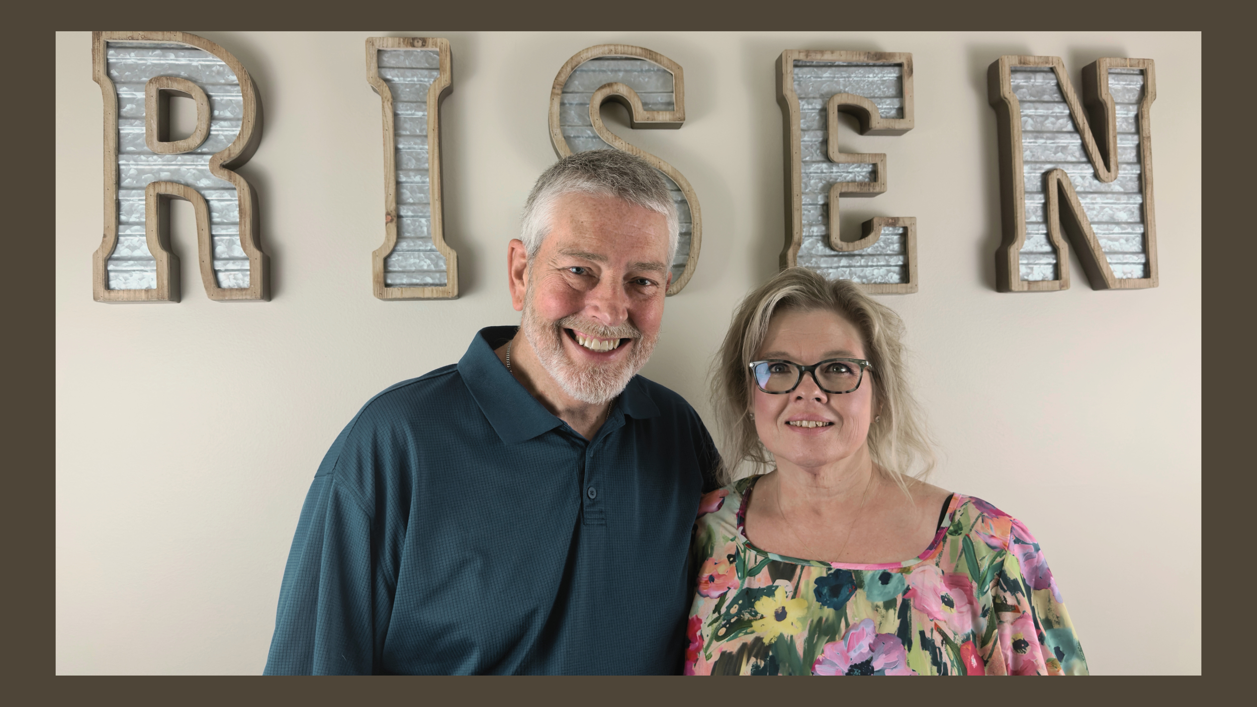 A smiling man and woman posing in front of a wall with large decorative letters spelling 'RISE'. The man has short gray hair and a beard, wearing a dark teal collared shirt. The woman has gray hair, glasses, and is wearing a colorful floral blouse.