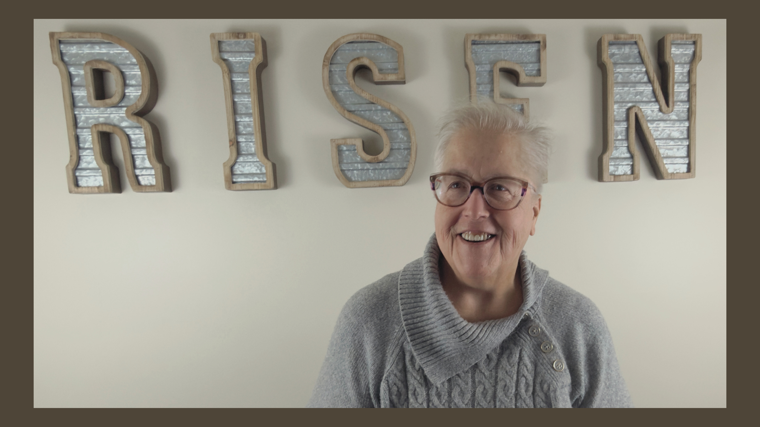 An elderly woman with short gray hair and glasses smiling in front of a wall with large decorative letters spelling NISEN.