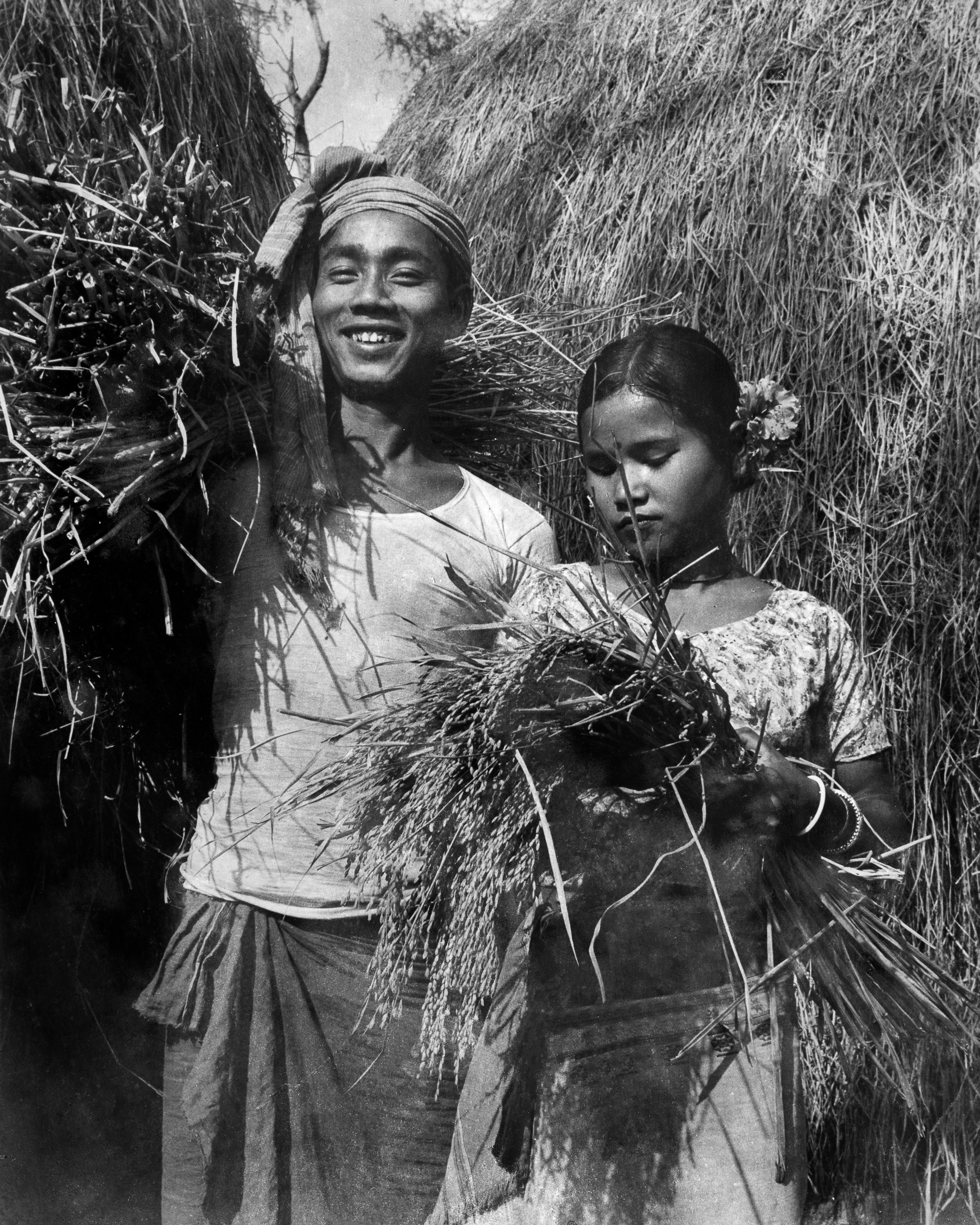 Mogh couple during harvest season with paddy collected from their rice fields. The woman wears a screen printed short blouse instead of a risha. Maitang or paddy is a common motif in Tripuri textiles. c.1977