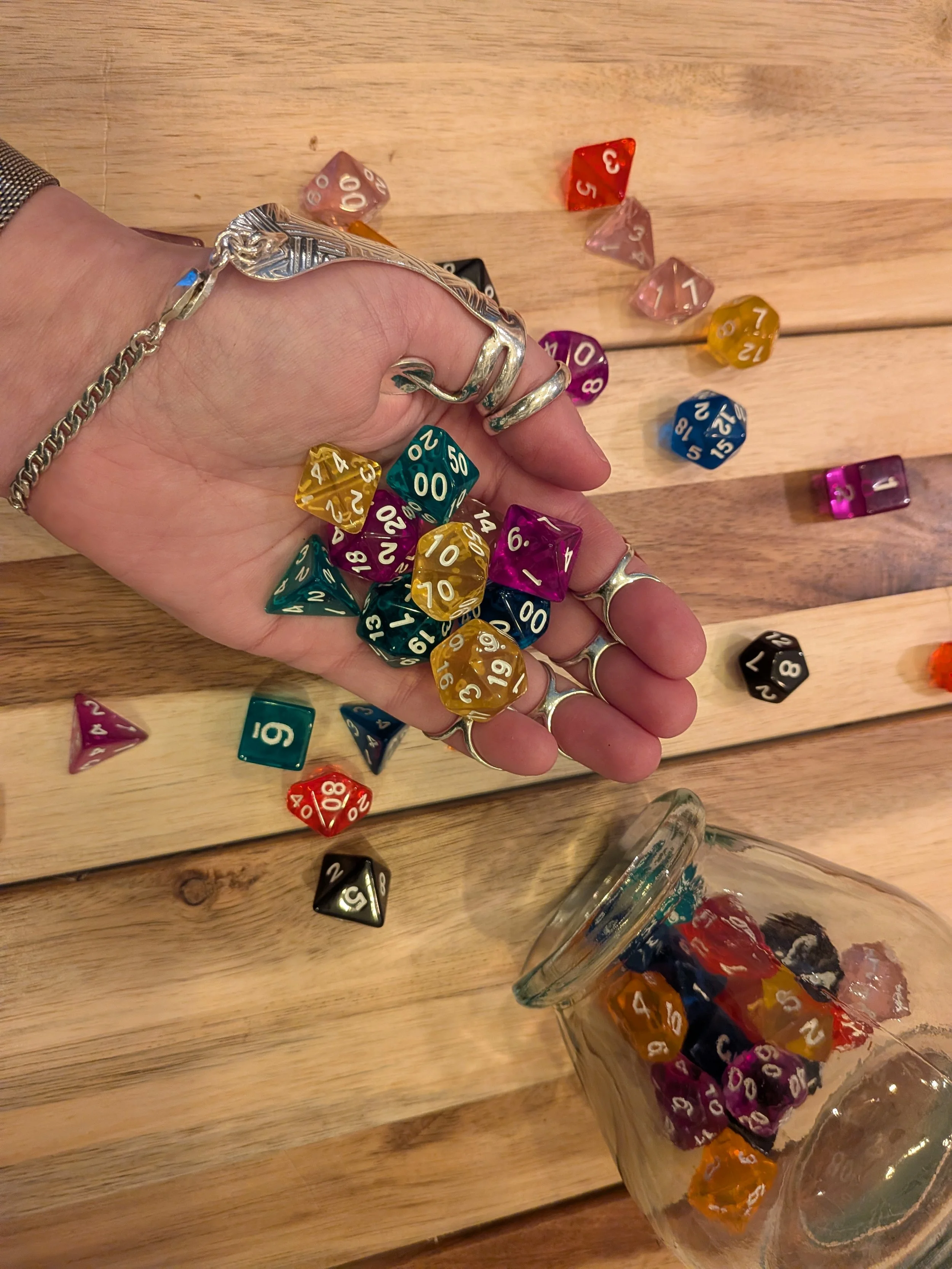 Nora's left hand (covered in silver hEDS splints from SIRIS) holds multicolored D&D dice, which also spill from an open glass jar onto a wood tabletop below