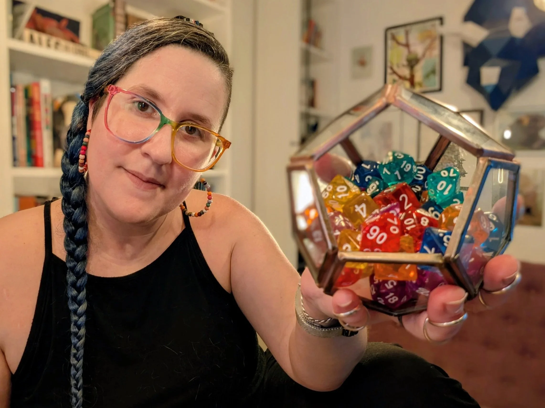 A woman with long blue hair, rainbow glasses, and rainbow beaded earrings holds a glass dodecahedron full of multicolor D&D dice