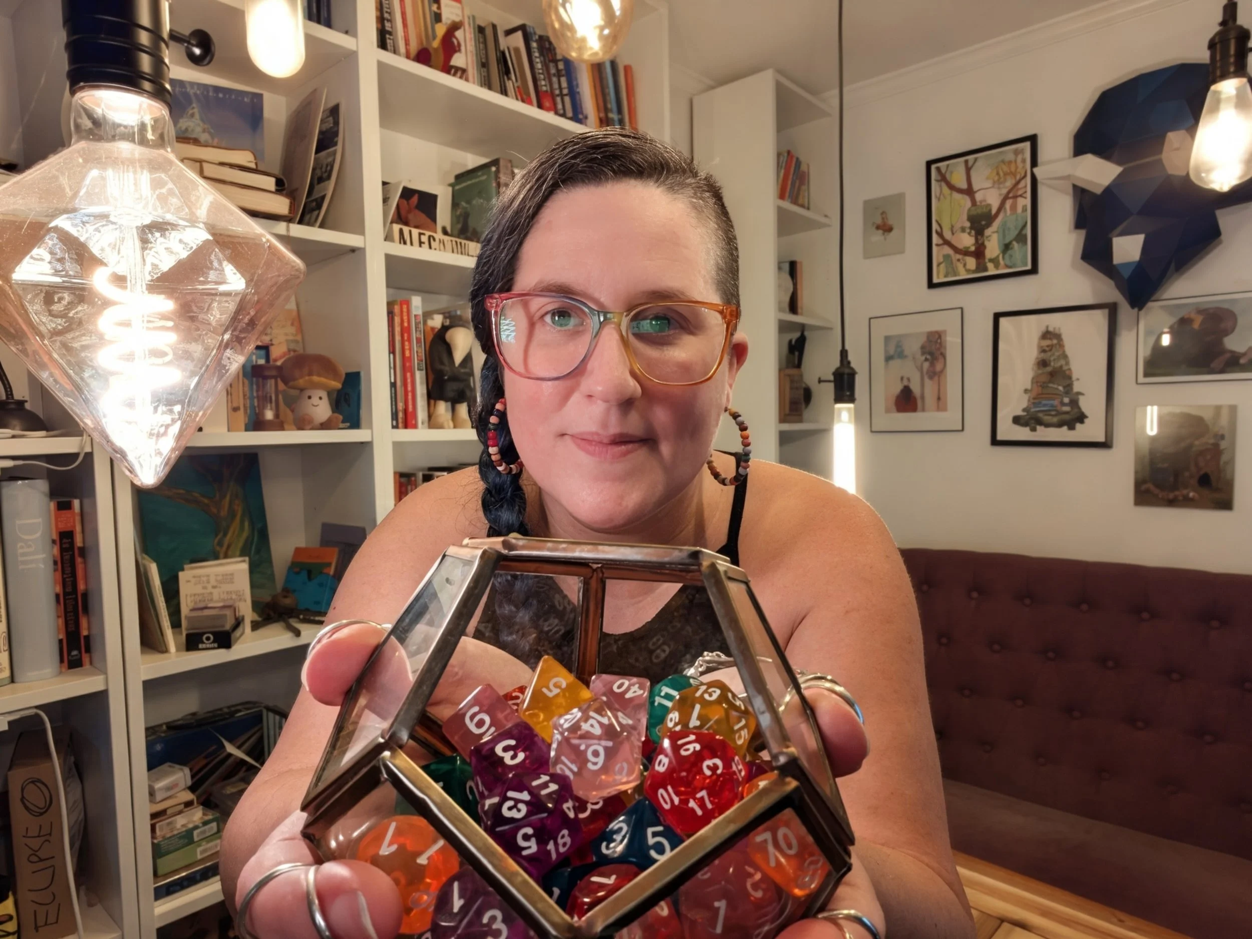 A woman with long blue hair, rainbow glasses, and rainbow beaded earrings holds a glass dodecahedron filled with rainbow D&D dice.  Behind her are bookcases and Edison-style bulbs in different shapes