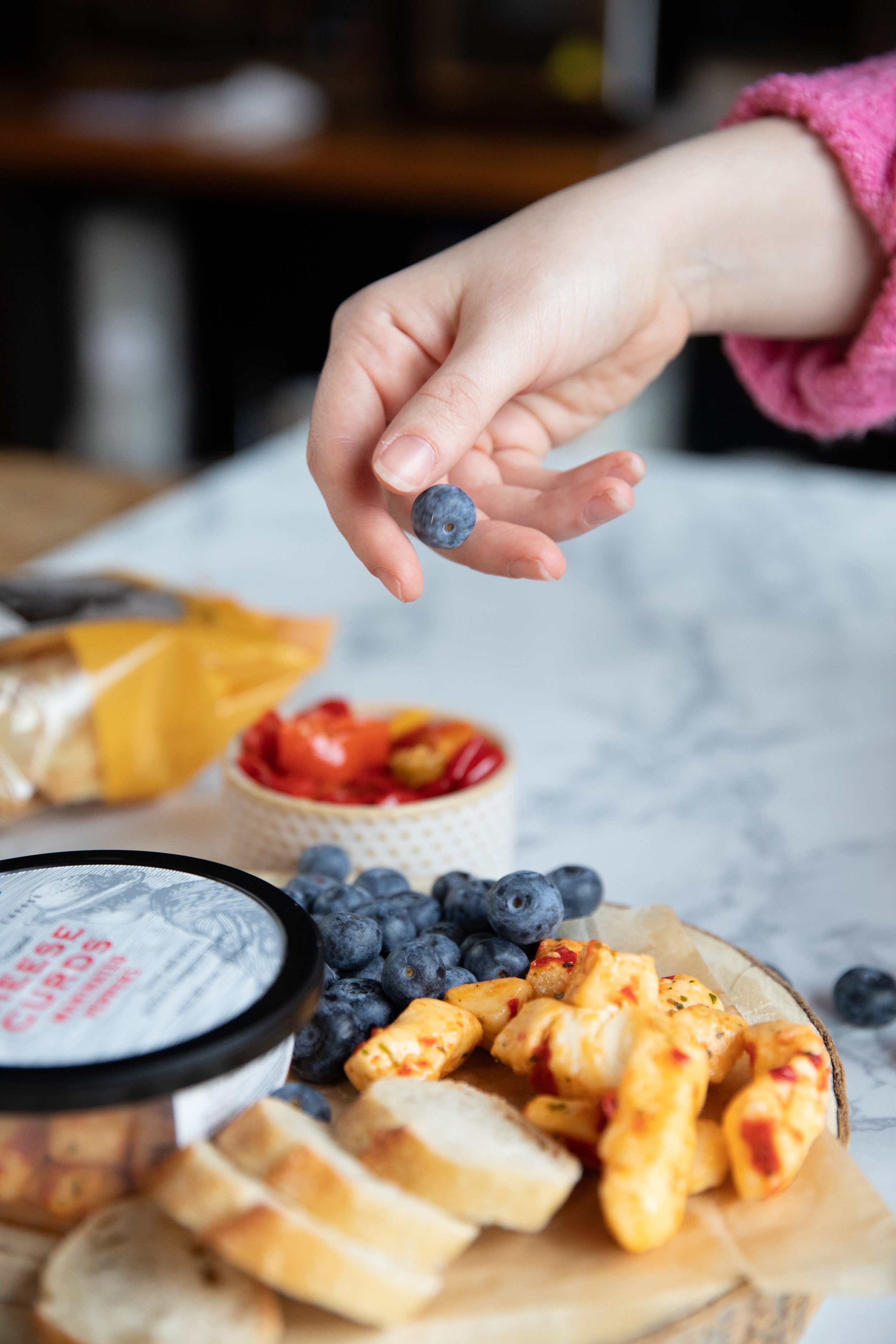 A hand holding a blueberry over a plate of food that includes sliced bread, blueberries, and pieces of baked chicken, with a container of spreadable cheese and small bowl of peppers in the background.