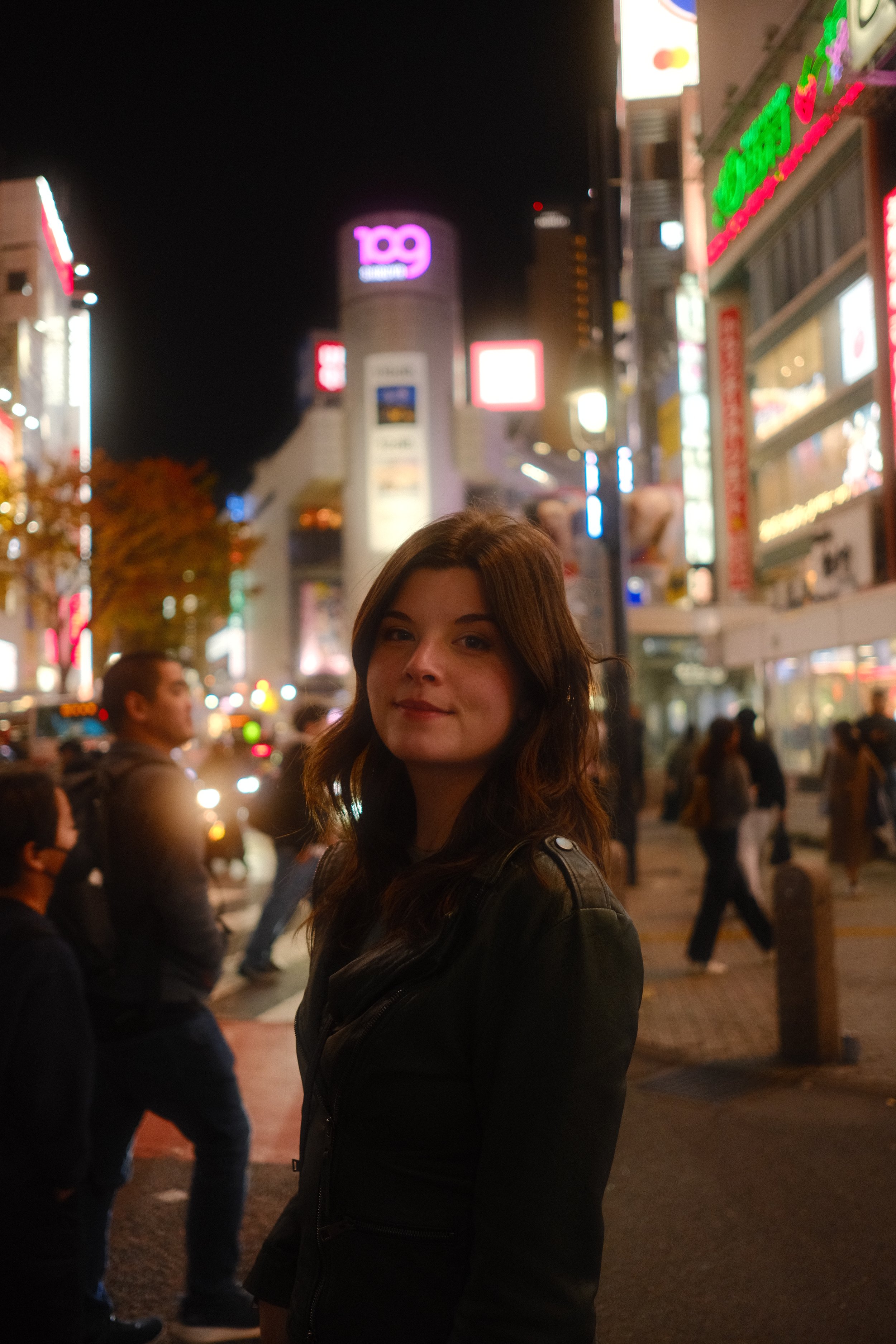 A young woman with brown hair stands on a city street at night, surrounded by illuminated signs and blurred pedestrians.