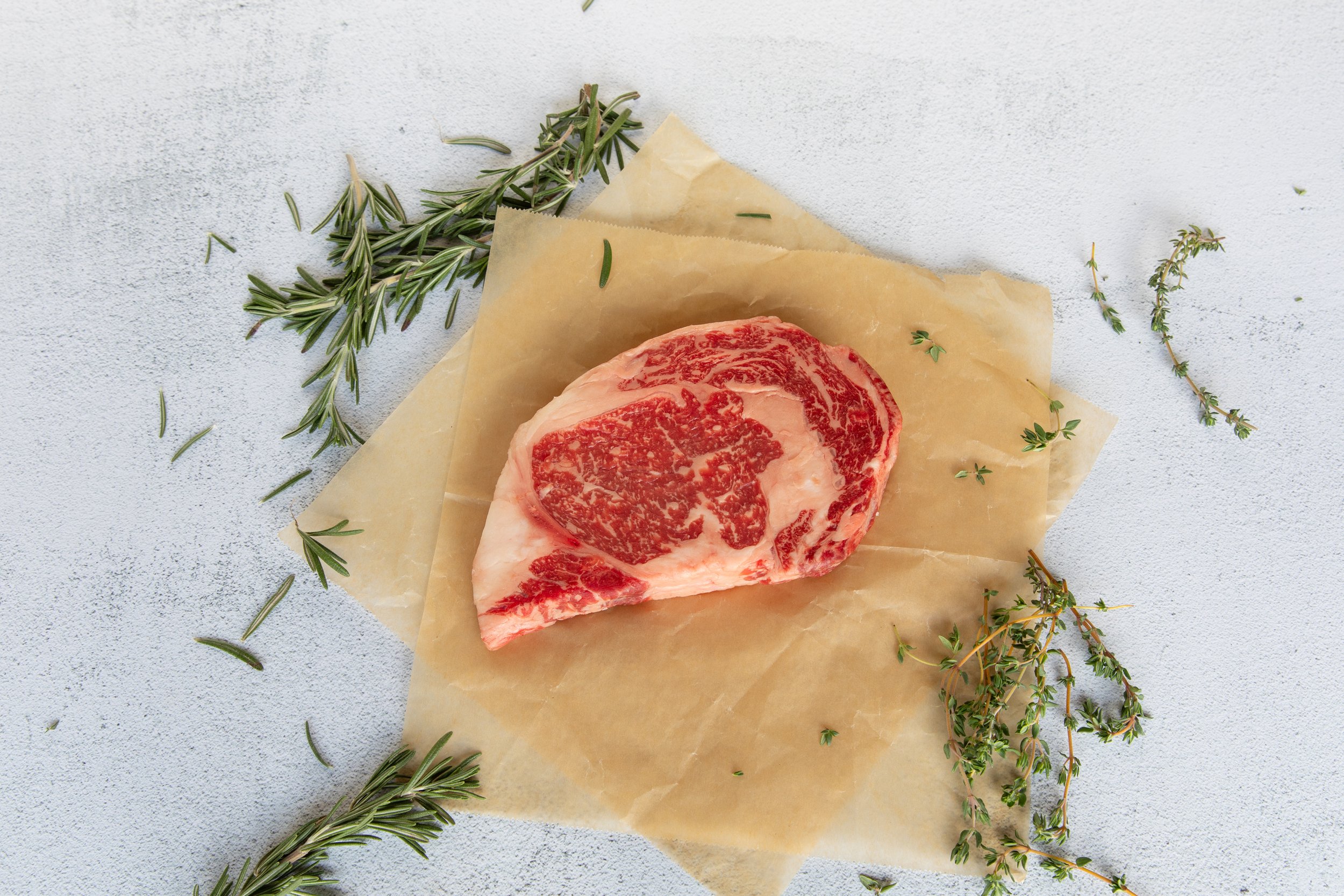 A raw, marbled steak on parchment paper, decorated with sprigs of fresh rosemary and thyme on a light gray textured surface.