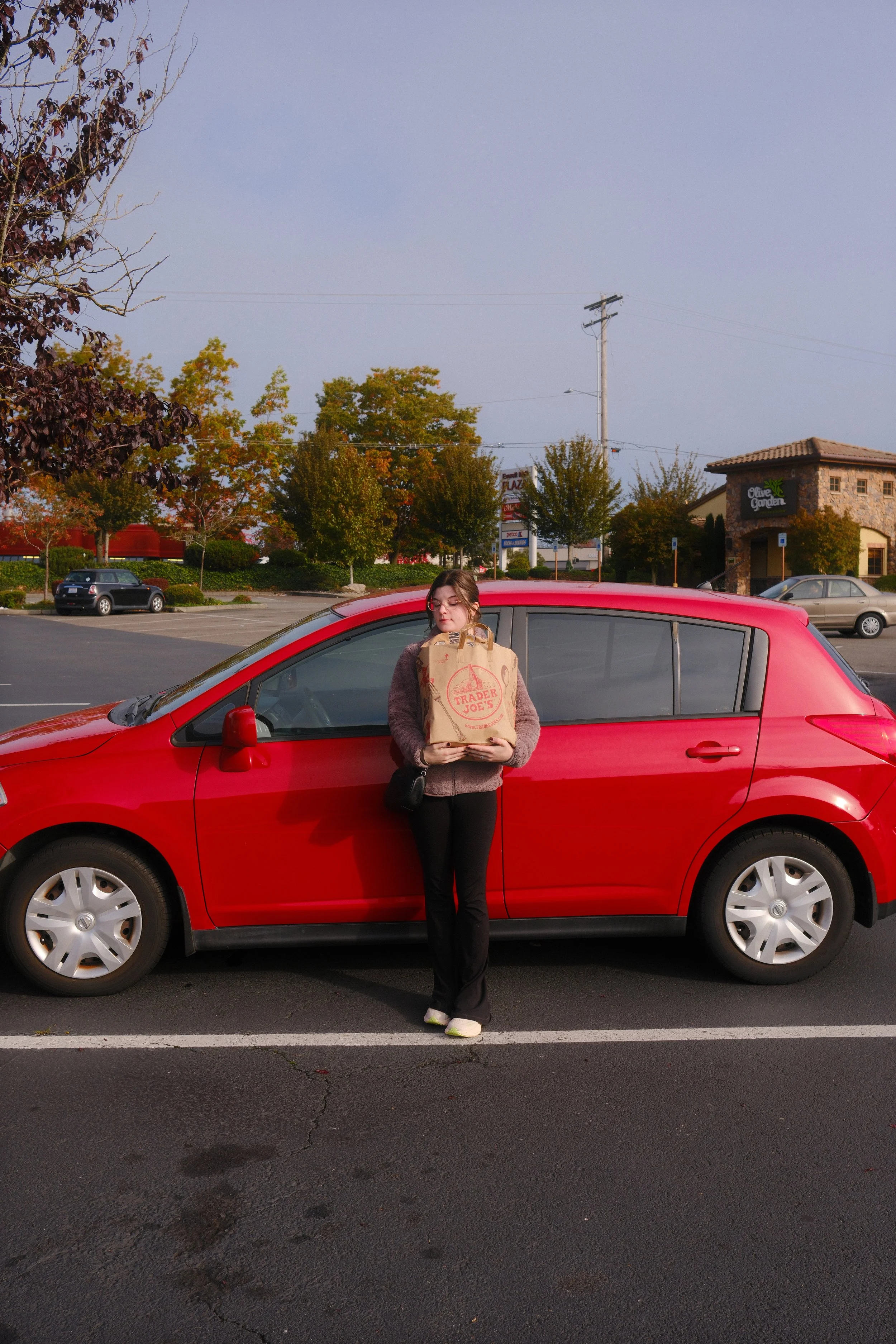 A woman standing in front of a red car holding a Trader Joe's shopping bag.