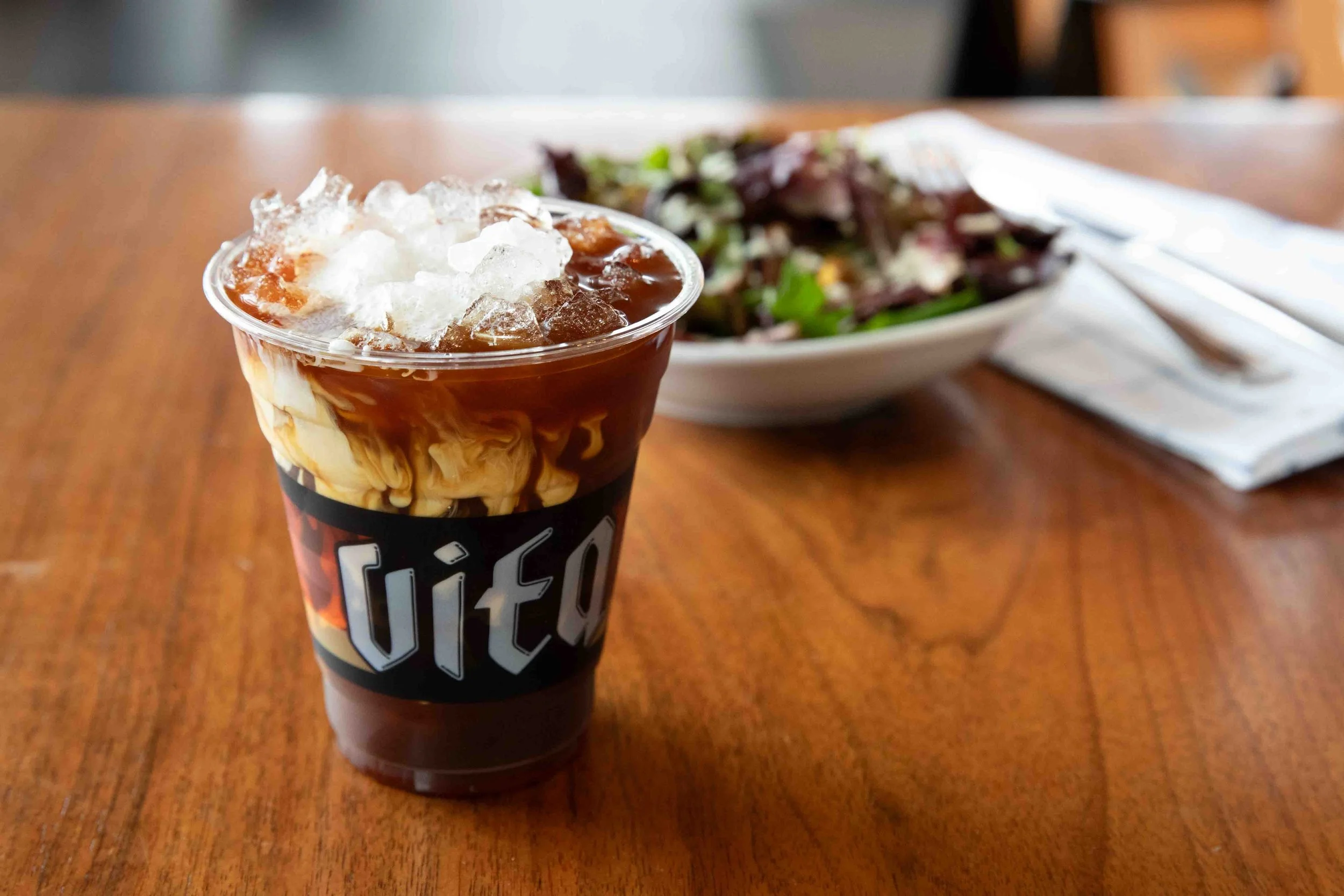 A plastic cup of iced coffee with cream and ice, placed on a wooden table next to a bowl of salad and napkin with utensils.