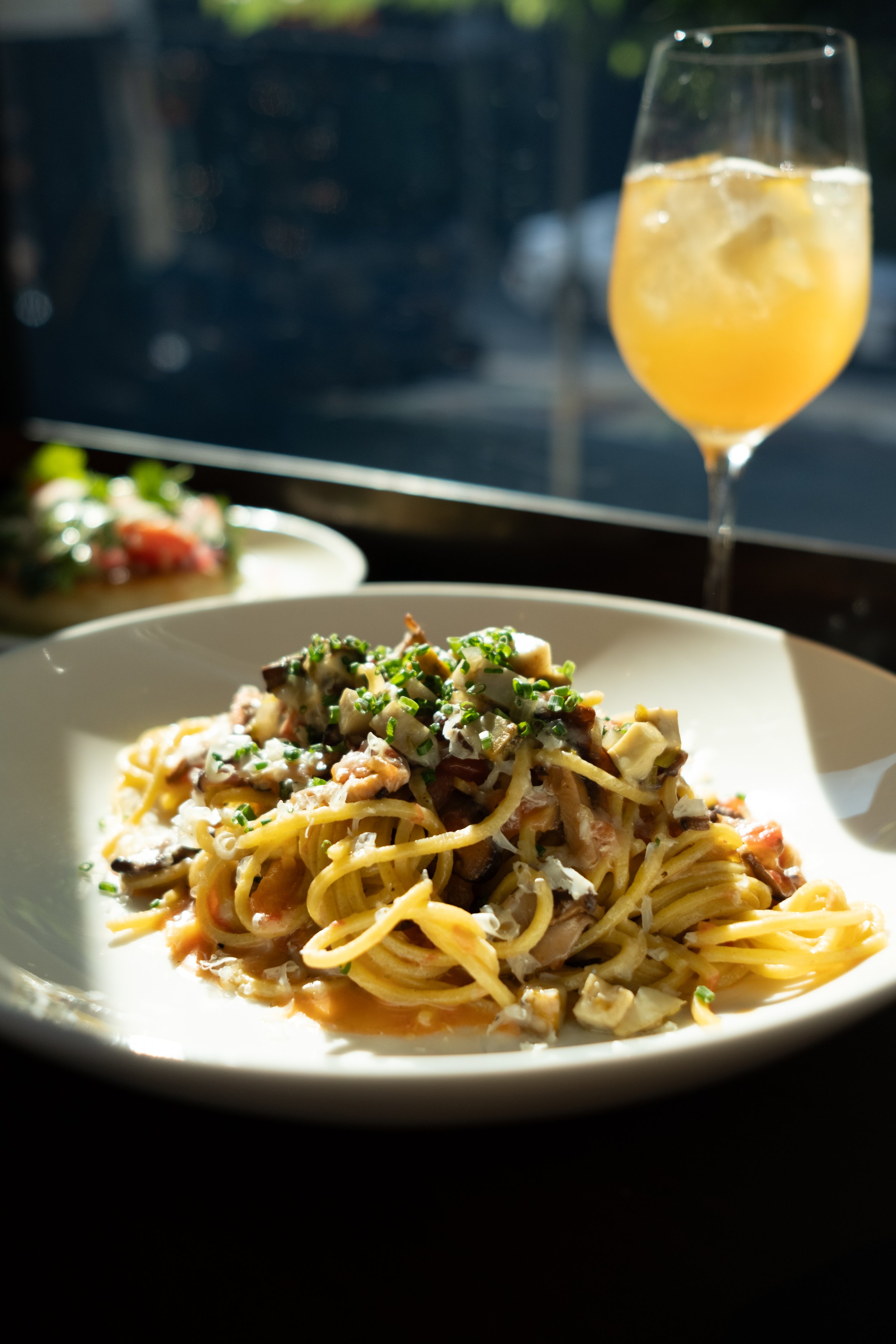 A plate of pasta with mushrooms, cheese, and herbs beside a glass of yellow beverage with ice, on a table near a window with sunlight.