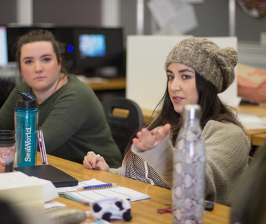 Two women sitting at a desk in a classroom, one wearing a gray knit hat and gesturing, the other listening with a water bottle and notebooks on the desk.
