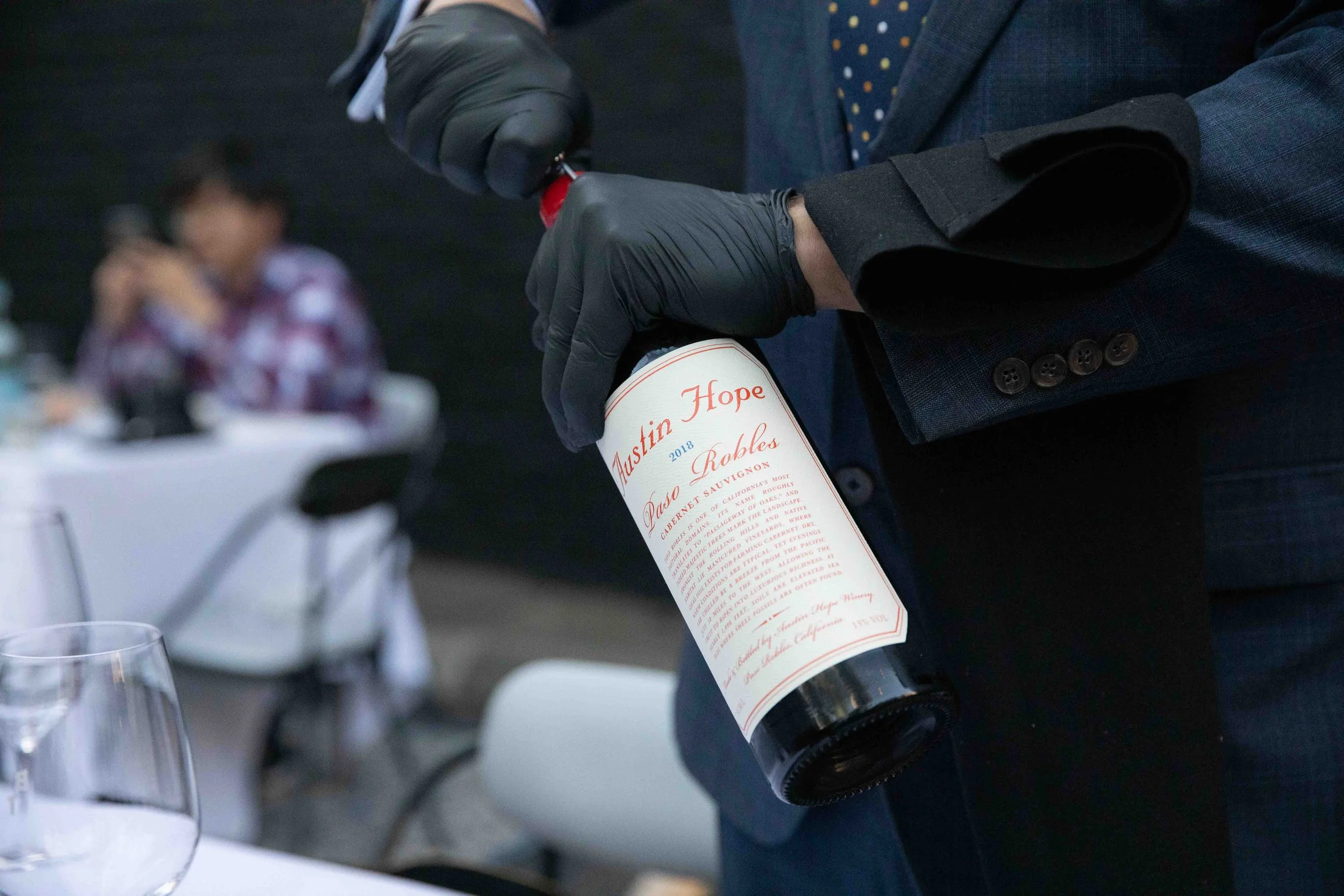 Man in suit opening a bottle of Austin Hope wine, with a blurred woman sitting at a table in the background.