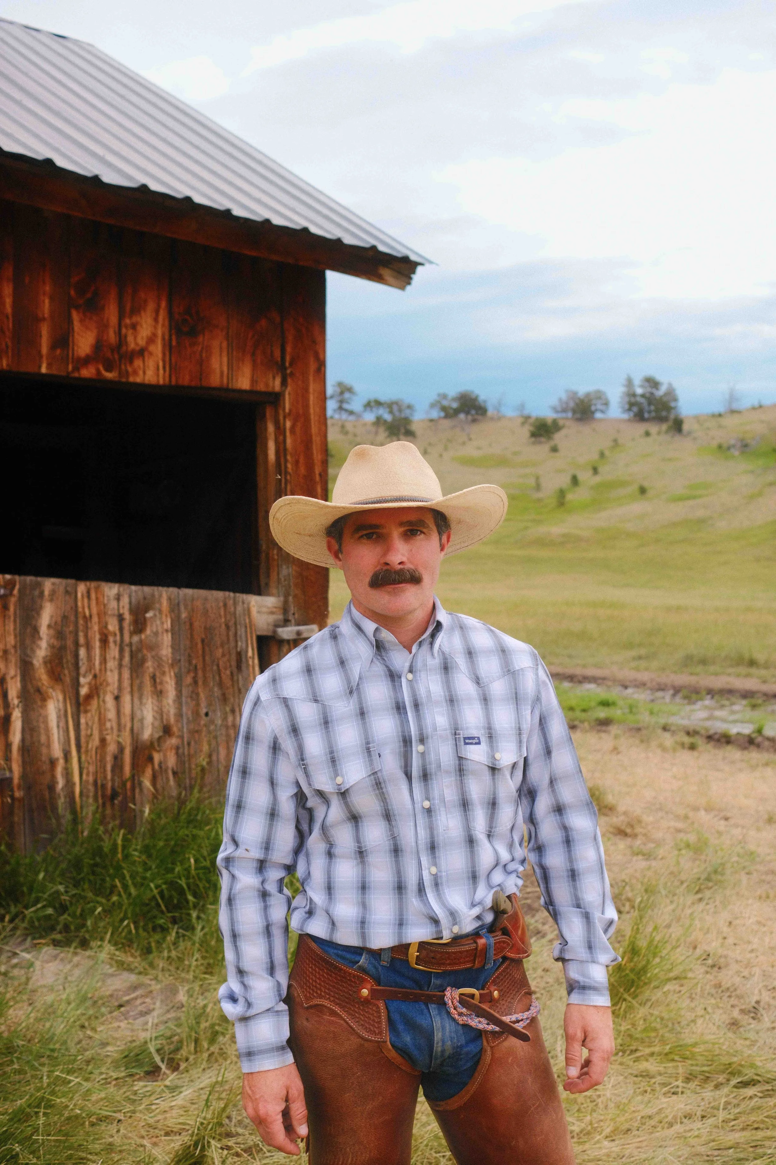 A man dressed as a cowboy standing in front of a rustic barn with a landscape of rolling hills and trees in the background. He is wearing a straw cowboy hat, a plaid button-up shirt, and brown leather chaps.