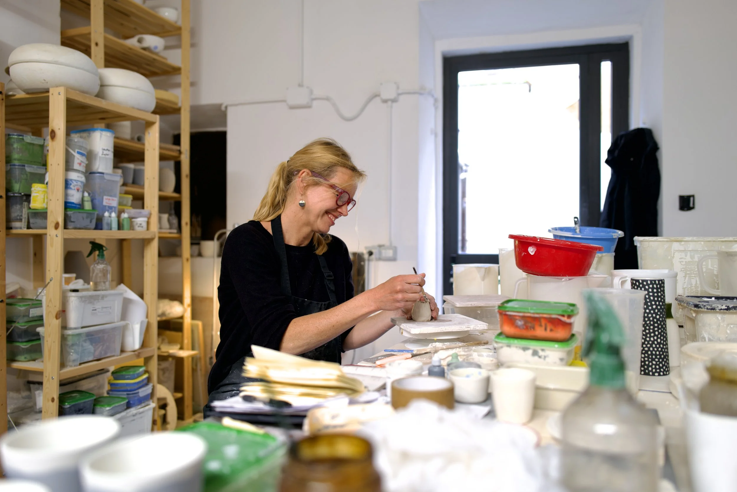 A woman with blonde hair and red glasses is working on a pottery piece in a ceramics studio surrounded by various bowls, containers, and art supplies.