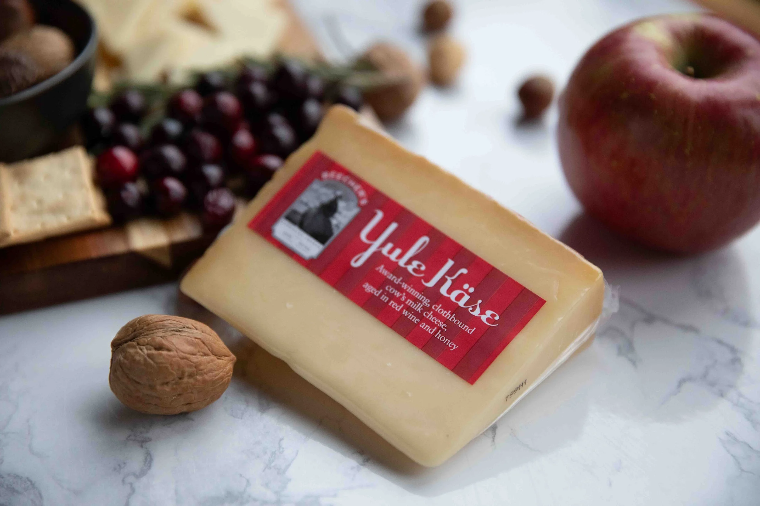 A block of Yale Kase cheese with red and white packaging, a walnut, an apple, grapes, and some cheese and crackers on a wooden board on a marble surface.