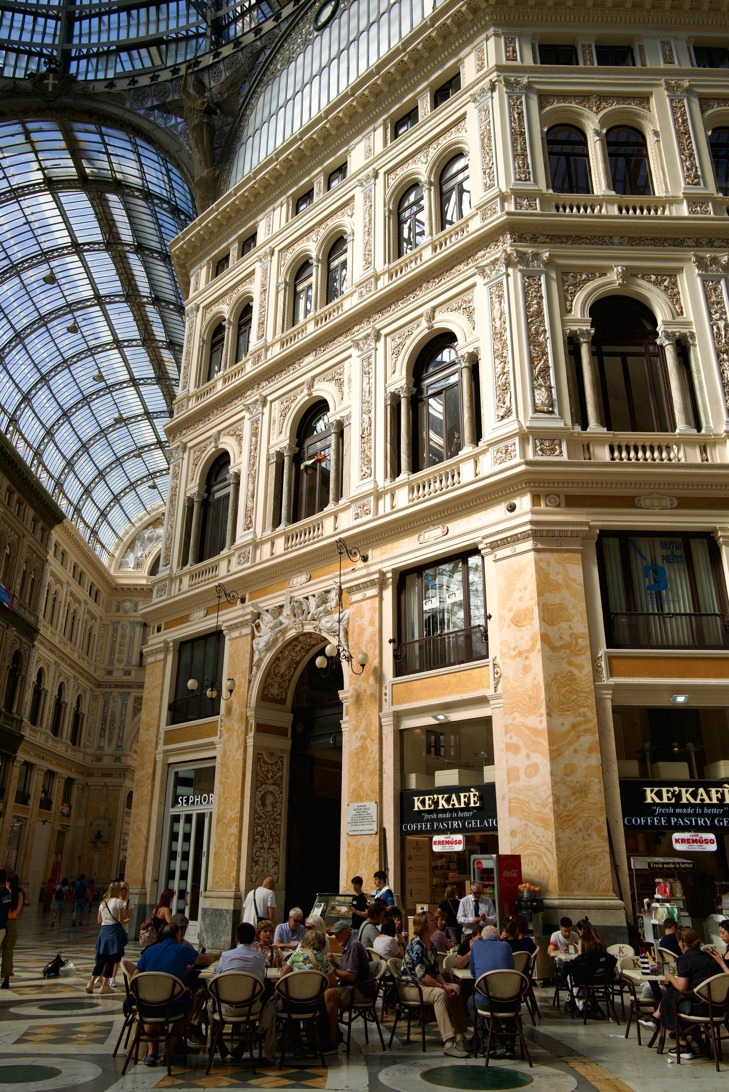 People sitting at outdoor tables in a shopping mall with ornate architecture and a glass ceiling.