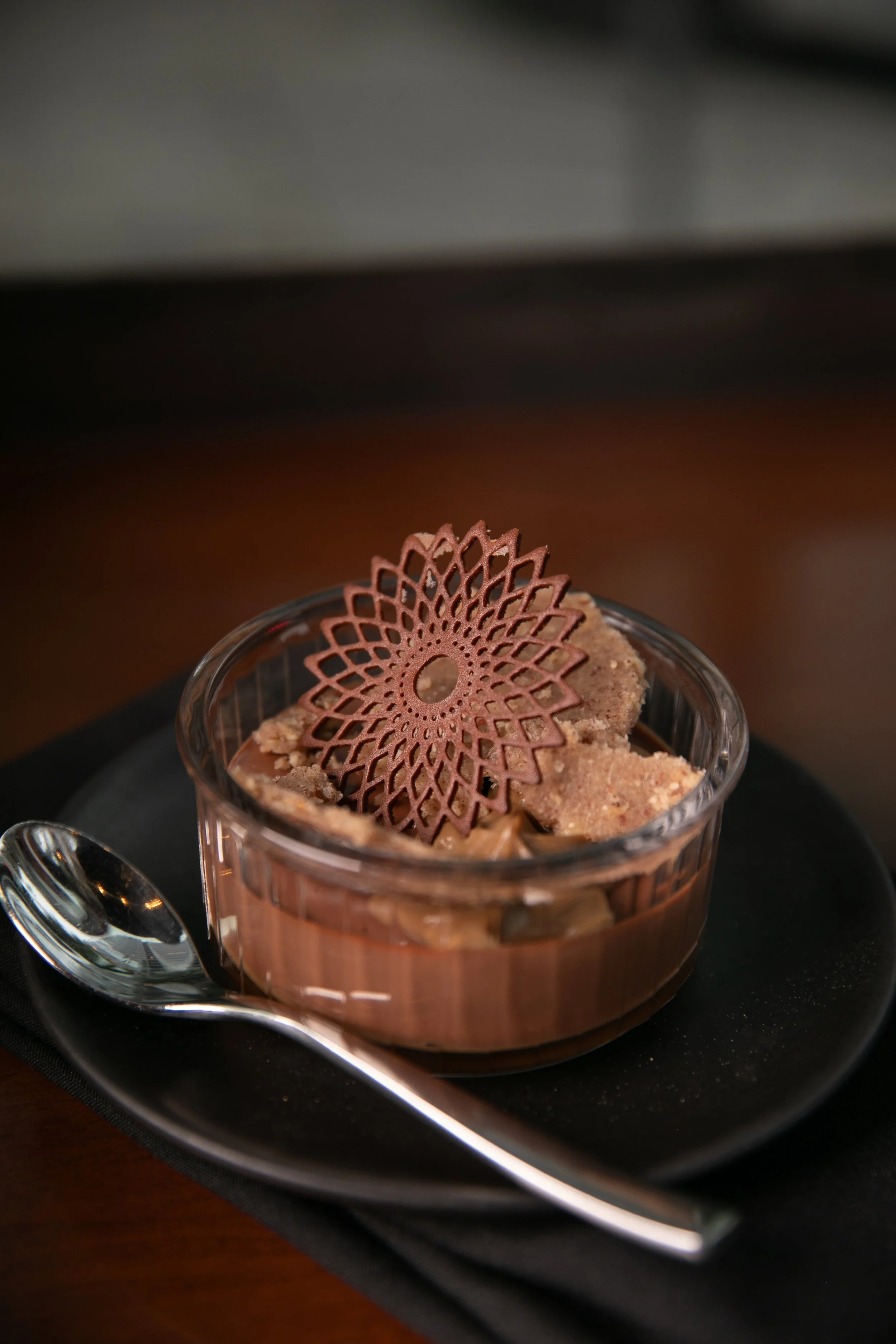 Chocolate dessert in a glass cup topped with a decorative chocolate wafer, served on a black plate with a spoon.