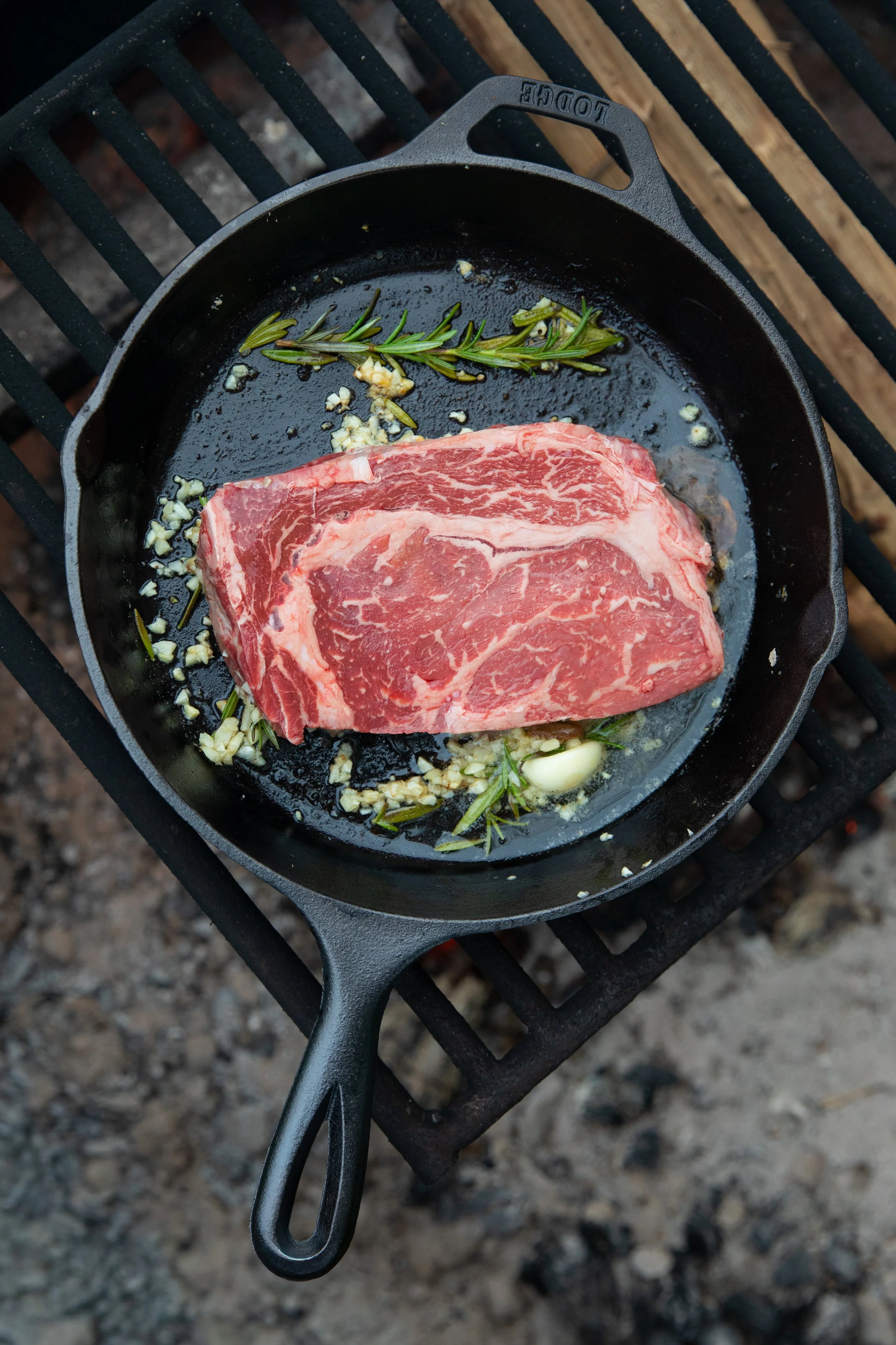 A cast iron skillet with raw marbled steak, garlic, rosemary, and minced garlic on a grill over charcoal.