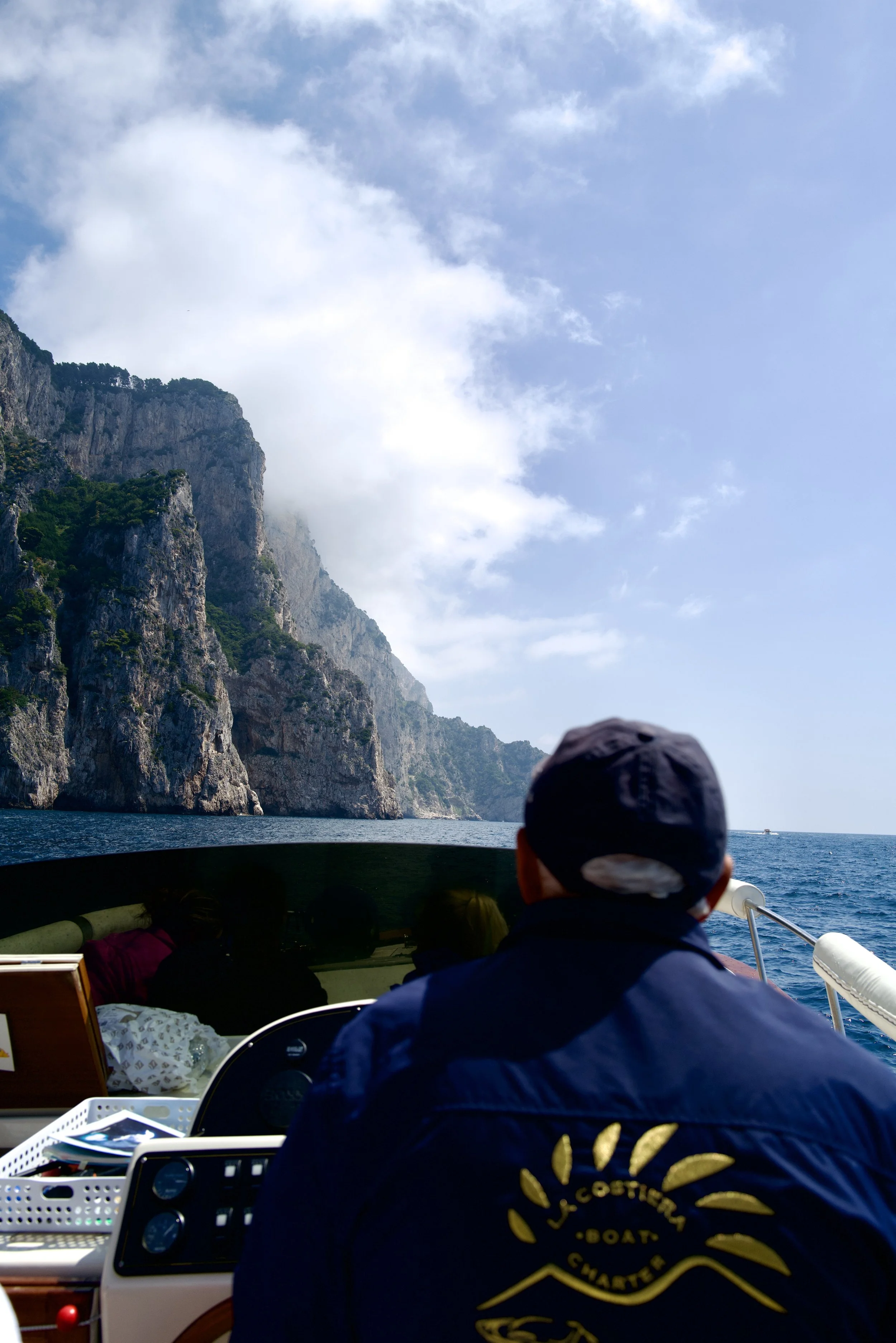 People on a boat tour near island cliffs, with one person in the foreground wearing a navy jacket that says 'L.COSTIGLIA BOAT CHARTER', facing the ocean and rocky cliffs under a partly cloudy sky.