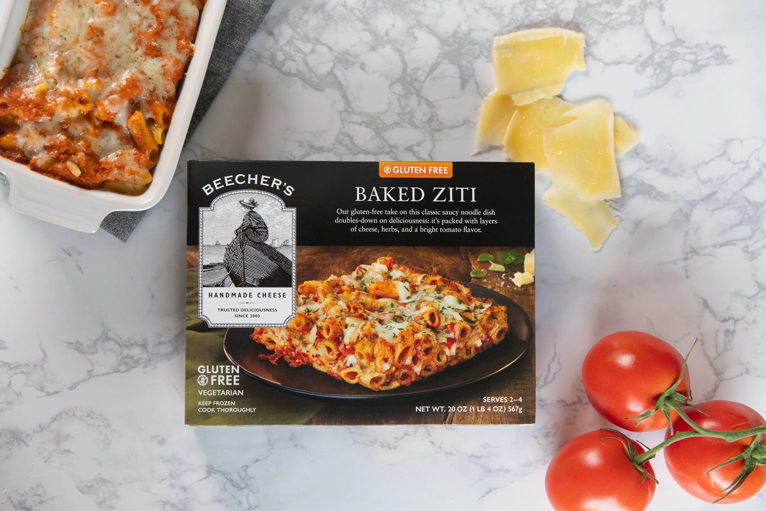 Package of Becher's Baked Ziti pasta with cheese, herbs, and tomato flavor on a white marble surface with three tomatoes and shredded cheese nearby.
