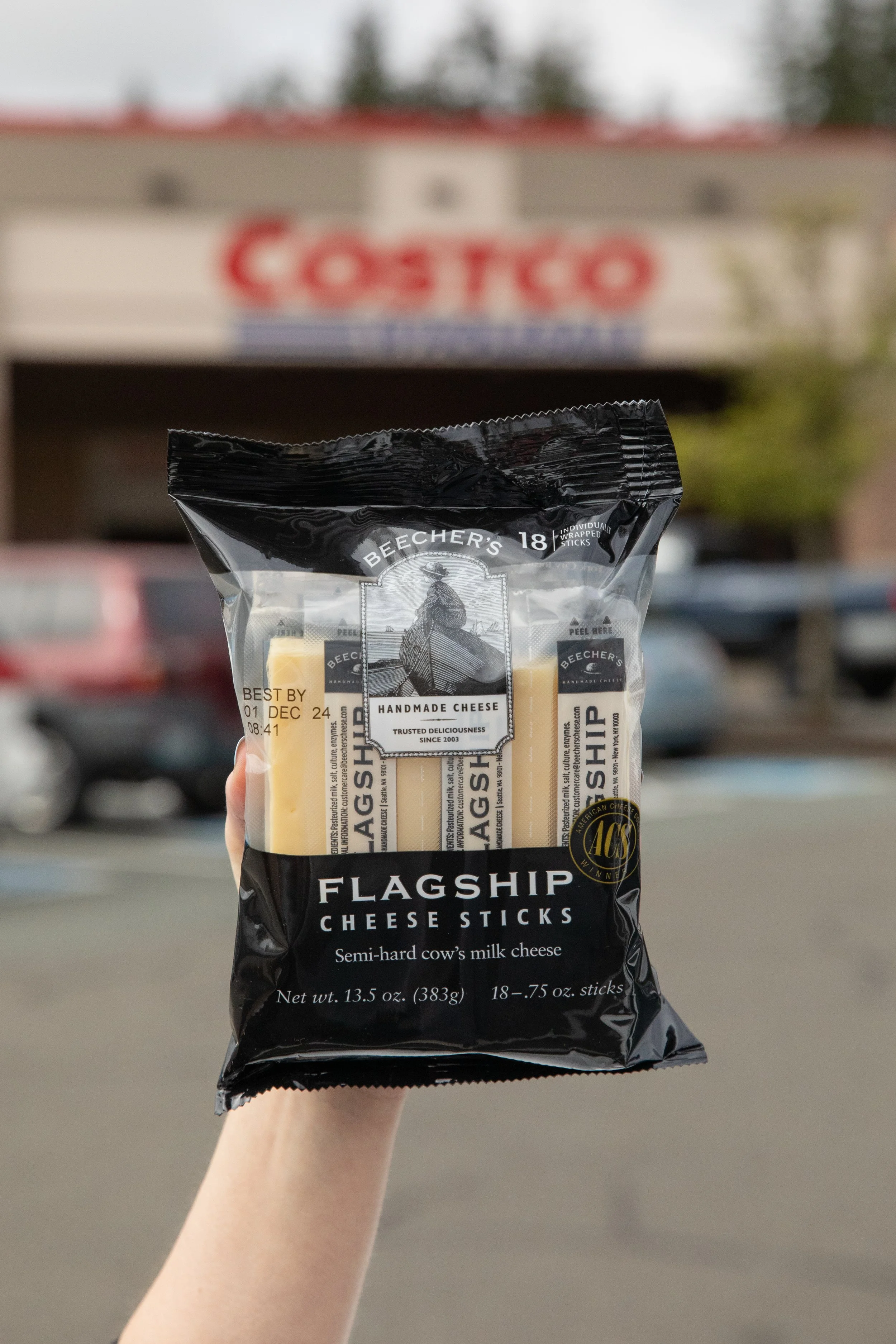 A hand holding a black package of Becher's Flagship Cheese Sticks in a parking lot outside a Costco store.