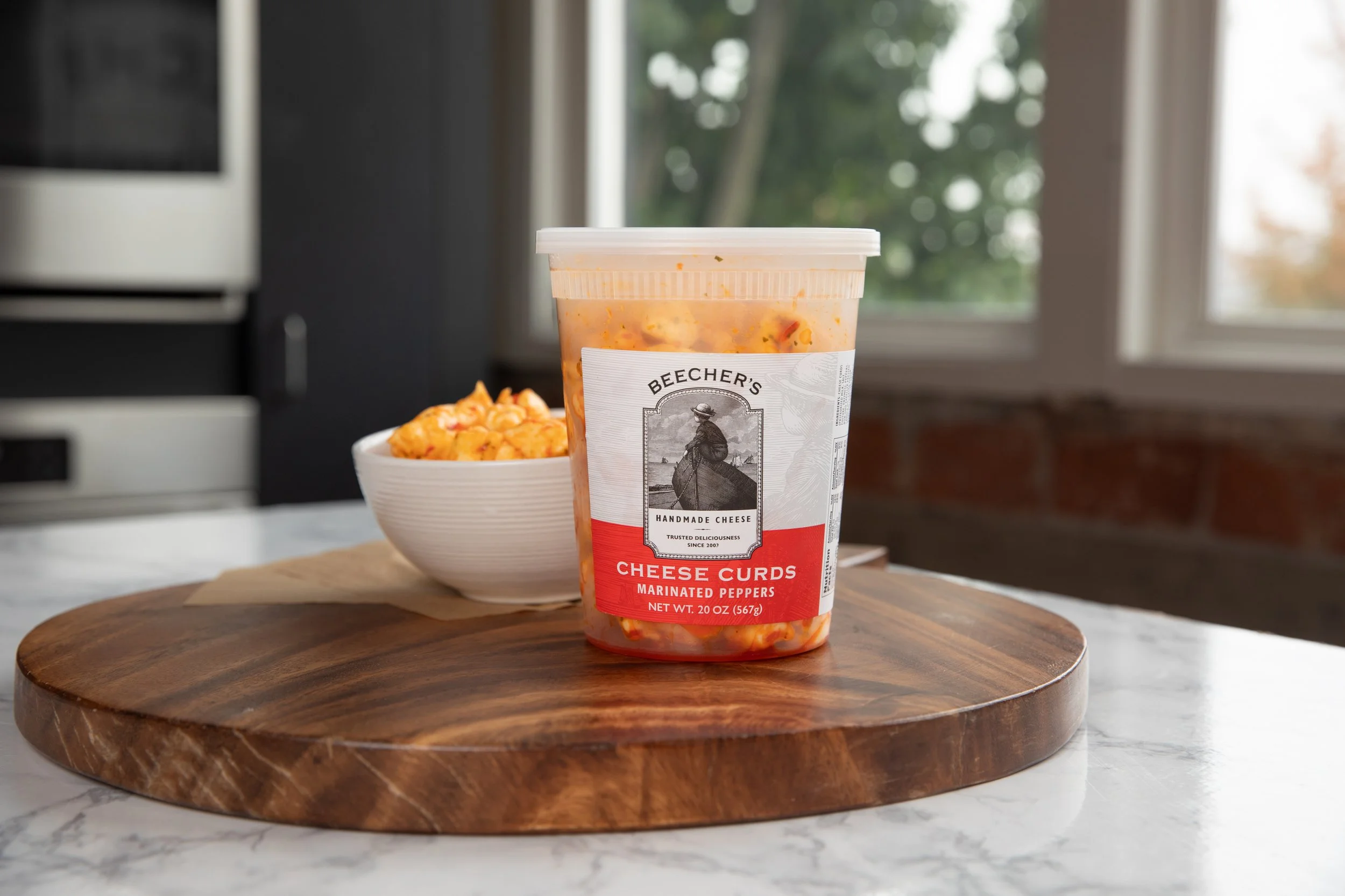 A container of Beecher's cheese curds with marinated peppers on a wooden board, with a bowl of cheese curds in the background in a kitchen setting.