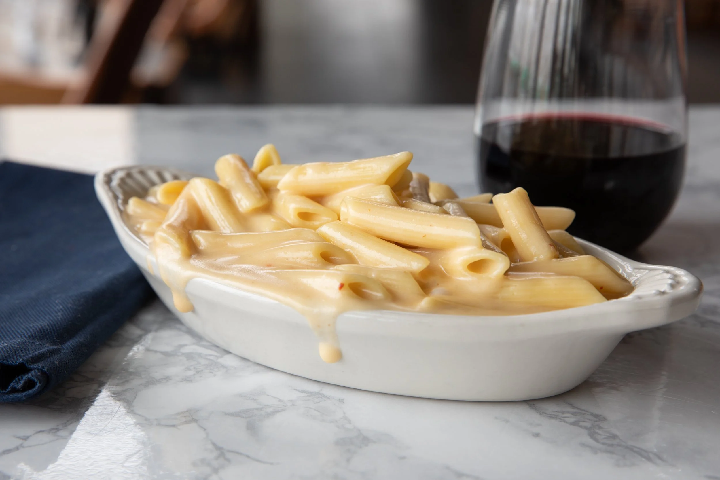 A white dish of cooked penne pasta with creamy cheese sauce, a glass of red wine, and a blue cloth napkin on a marble table.