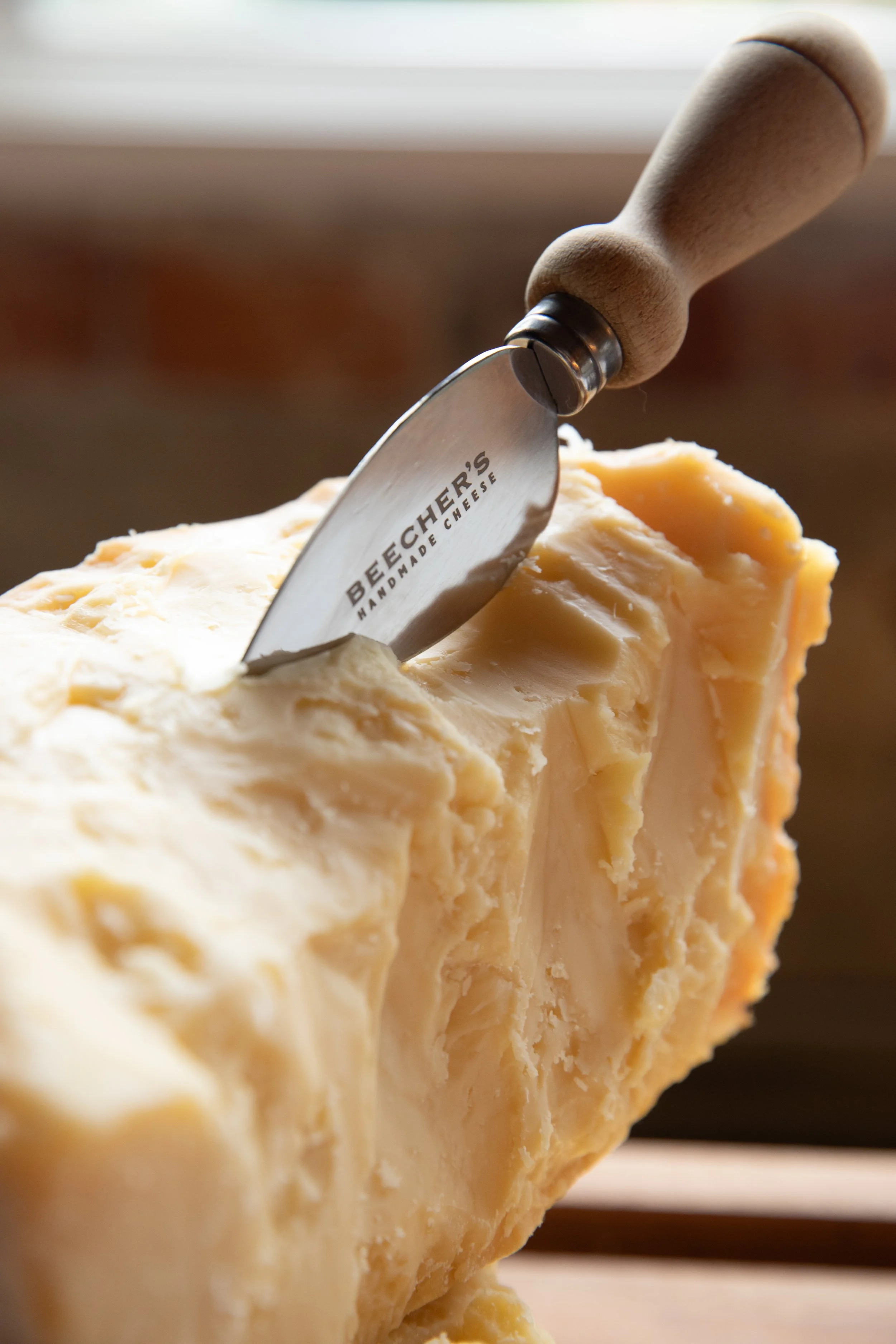 A wedge of cheese with a cheese cutter inserted into it. The cutter has a wooden handle and the blade is engraved with the words 'Beecher's Handmade Cheese.'