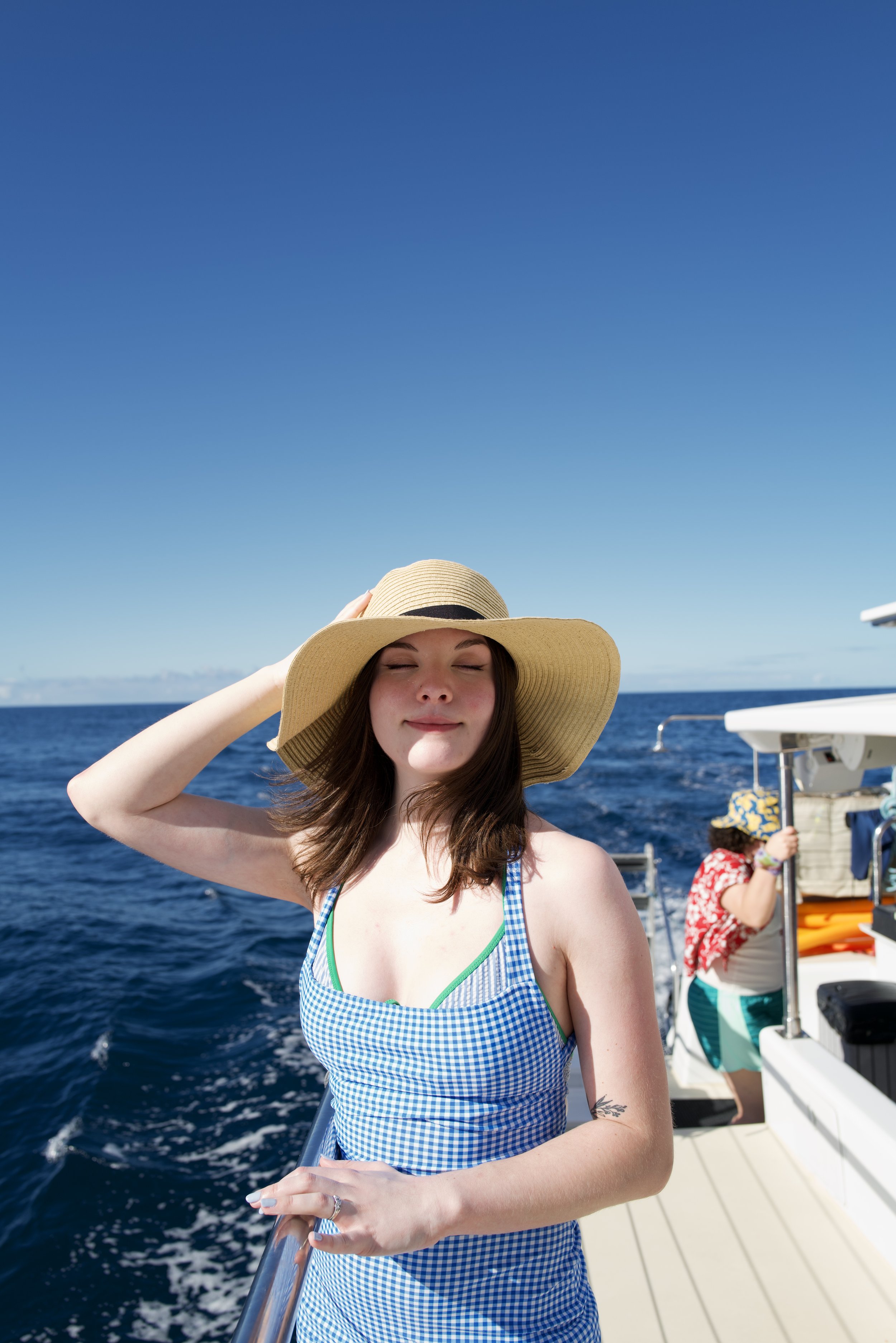 Young woman in a blue and white gingham dress with a large straw hat smiling with eyes closed on a boat, holding the railing, with the ocean and a clear blue sky in the background.