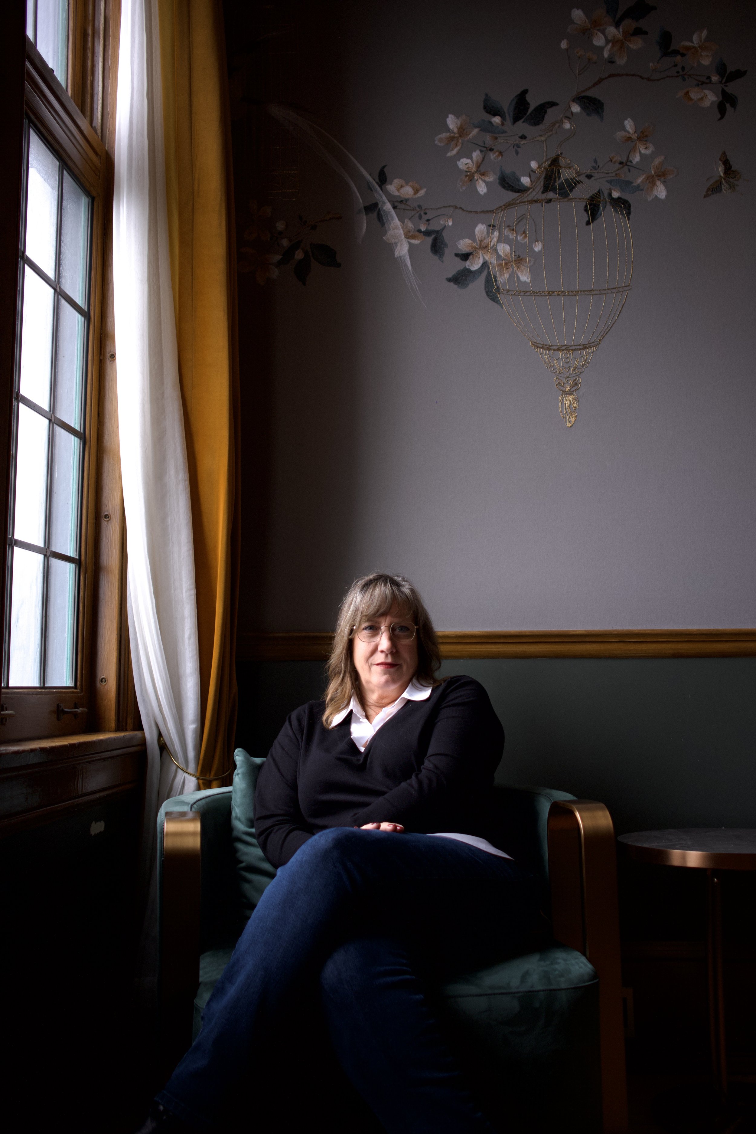 A woman sitting in a dimly lit room near a large window with yellow and white curtains, with floral and bird decor on the wall and a chandelier hanging from above.