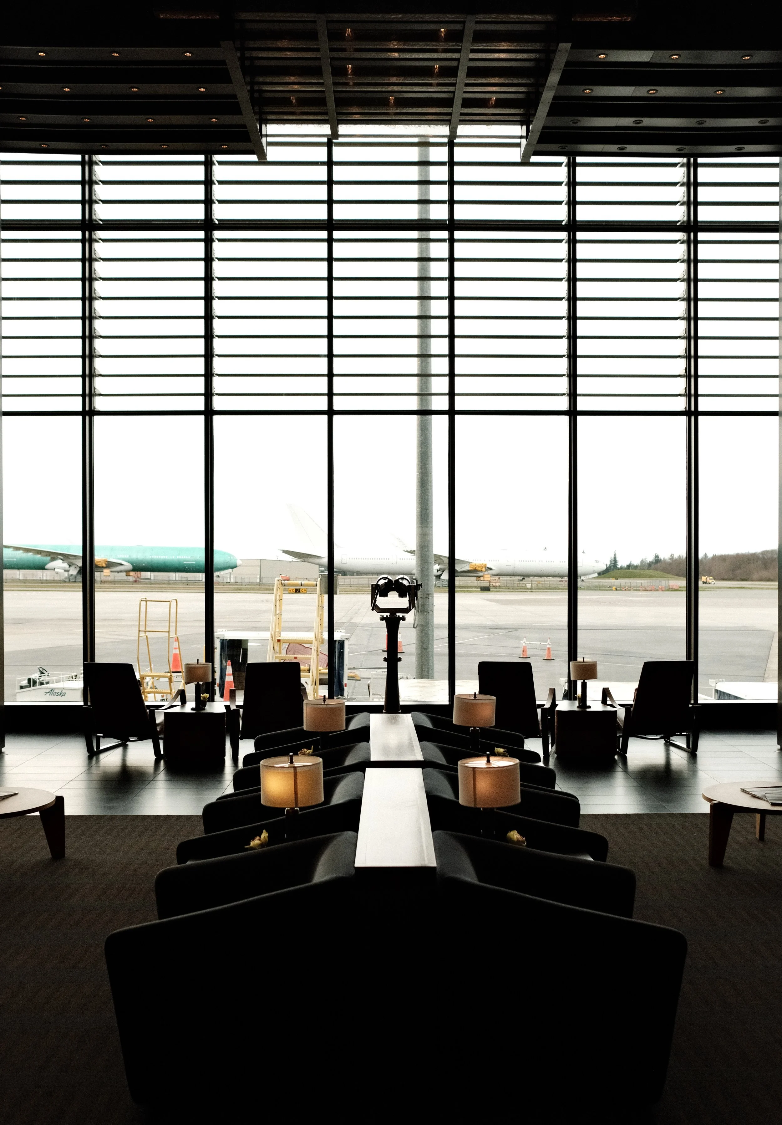 View of an airport terminal interior with a large window looking out onto other airplanes on the tarmac. Inside, there are chairs, small tables with lamps, and a telescope-like device facing the window.