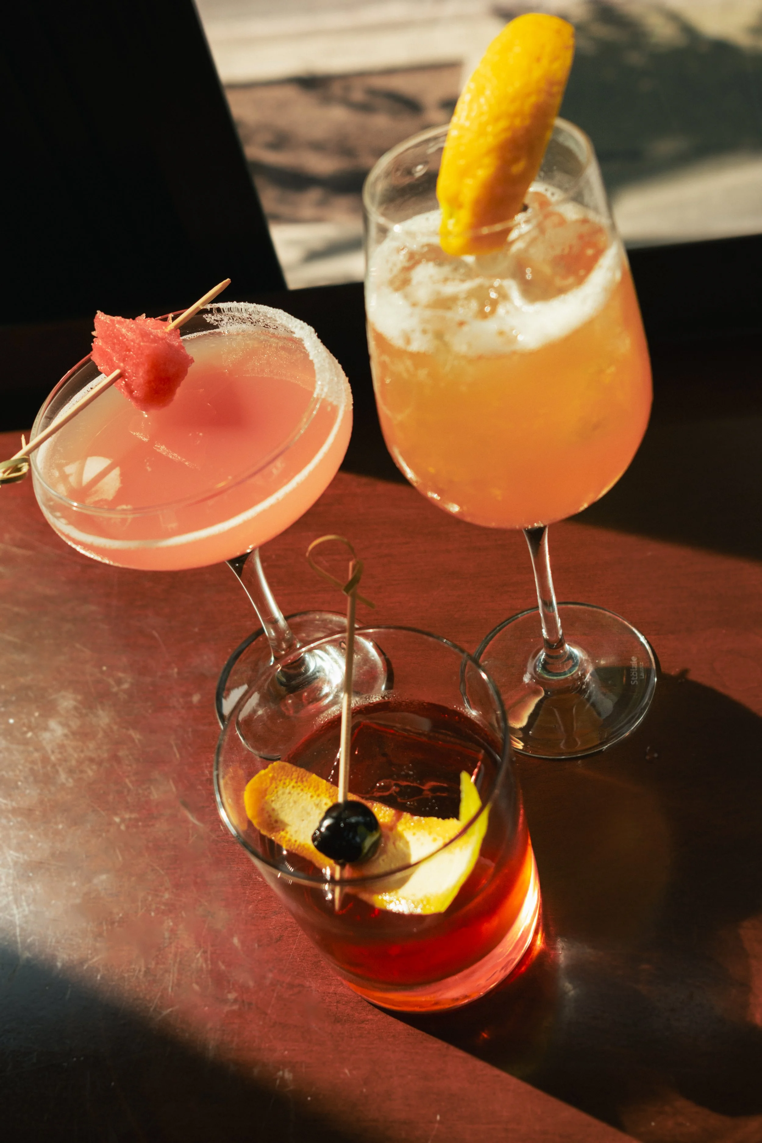 Three cocktails on a dark wooden table with a shadowed background. One pink cocktail with a pink garnish on a skewer, one yellow-orange cocktail with a lemon wedge, and a dark red cocktail with a lemon wedge and a cherry.