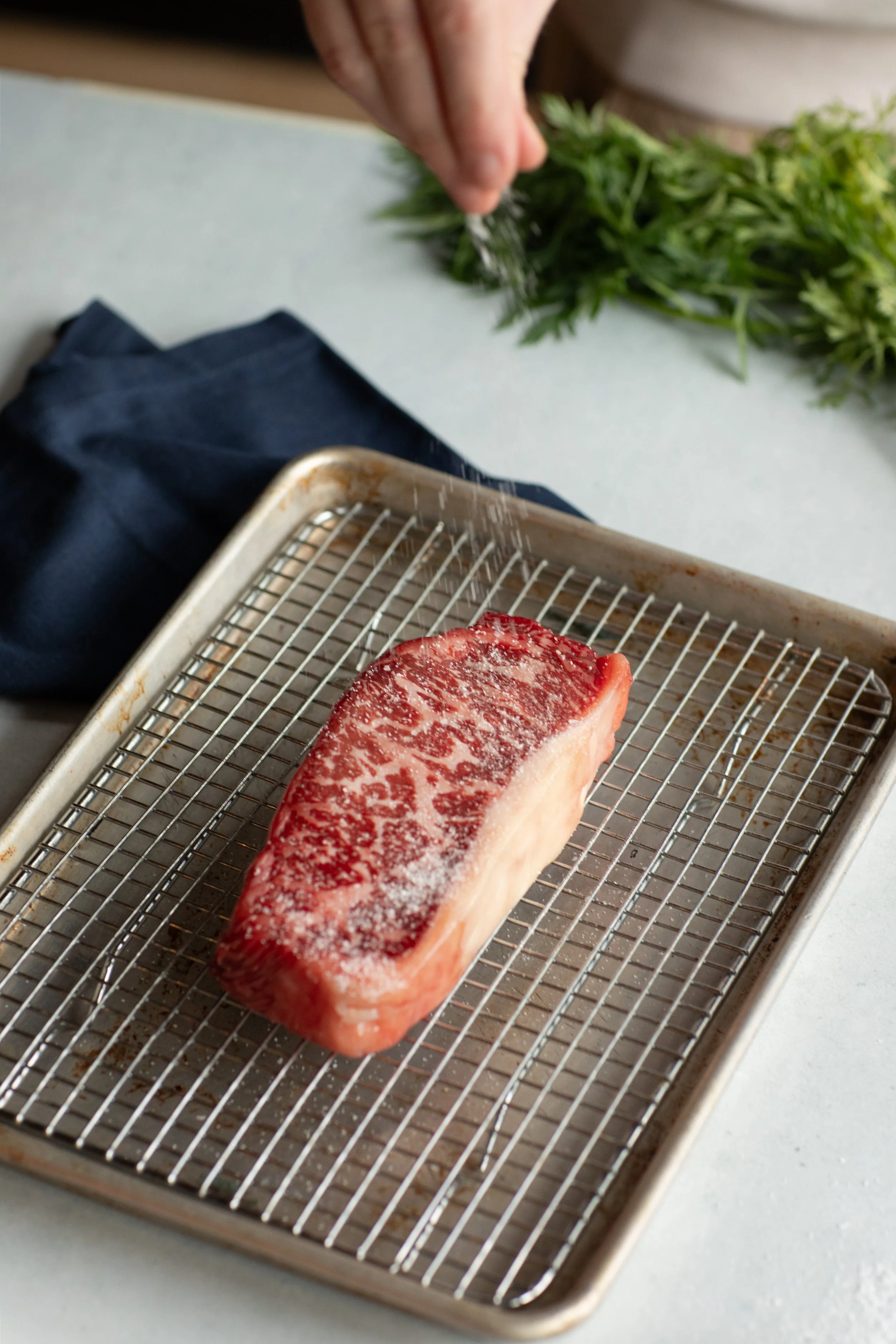 A piece of marbled beef resting on a metal wire rack on a baking sheet, with a person's hand sprinkling salt and herbs onto chopped greens in the background.