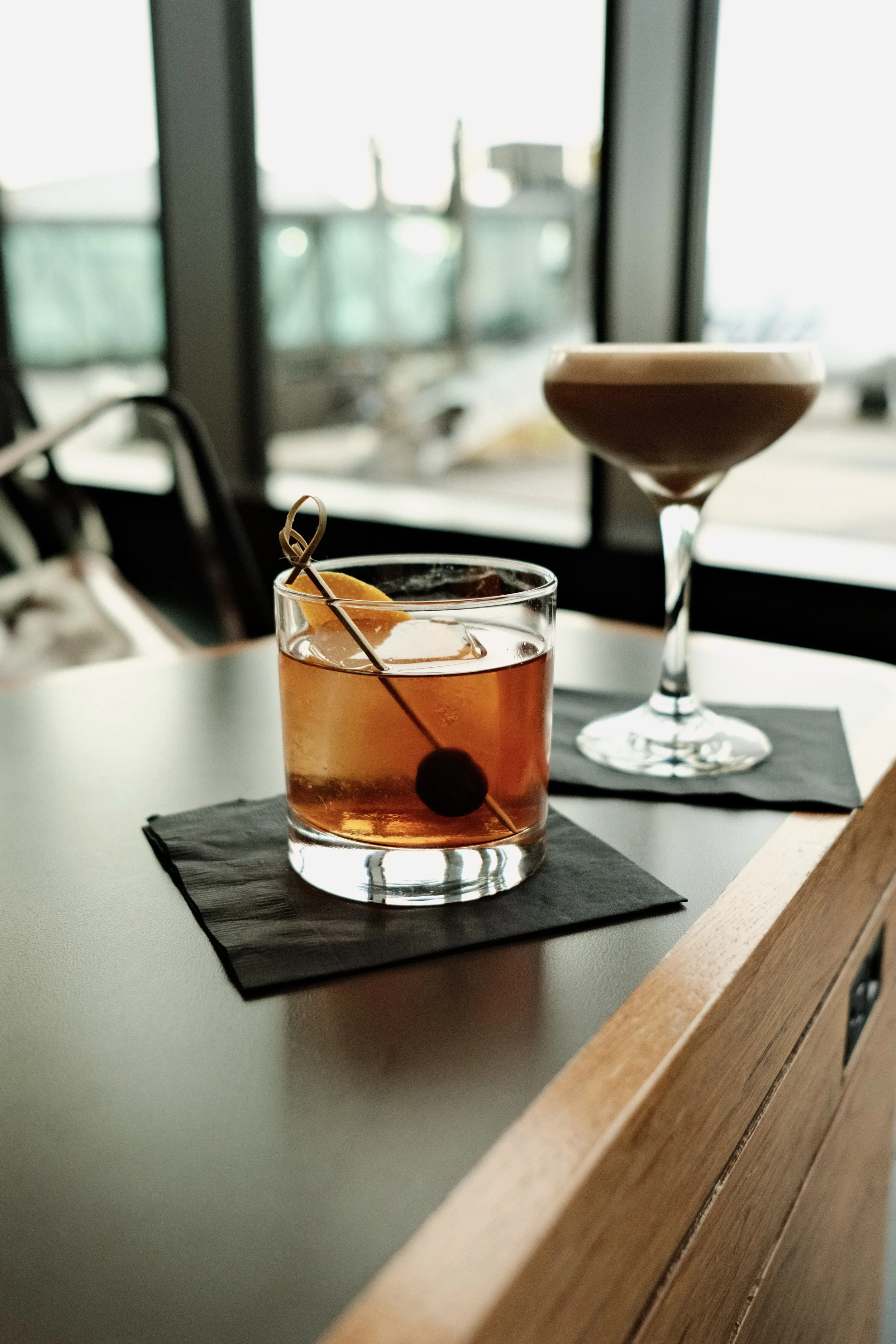Two cocktails on a table: one Old Fashioned with an orange twist and a cherry, and one in a coupe glass, set in front of large windows overlooking an outdoor area.