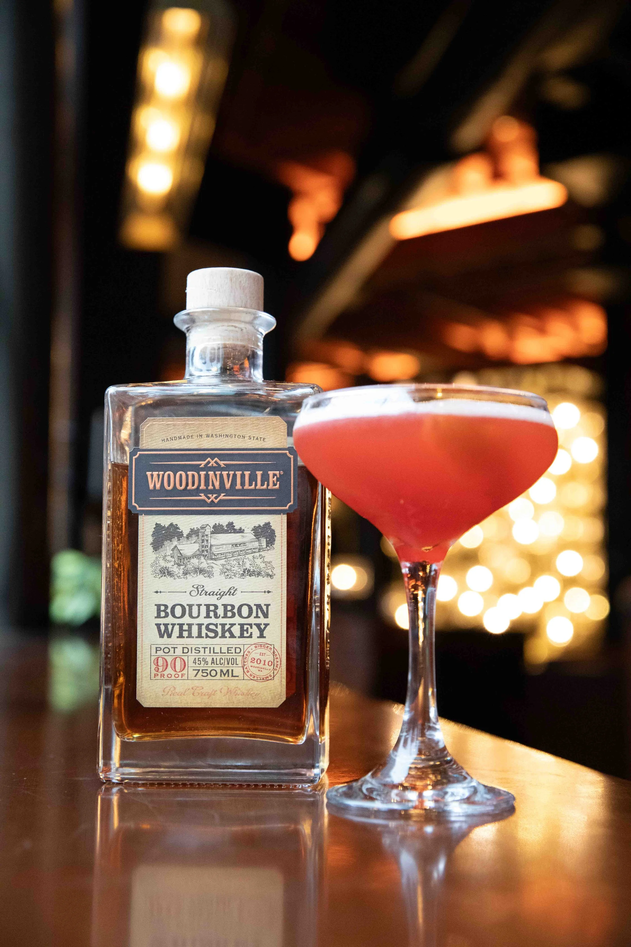 A bottle of Woodinville bourbon whiskey and a pink cocktail in a coupe glass on a bar counter with blurred warm lights in the background.