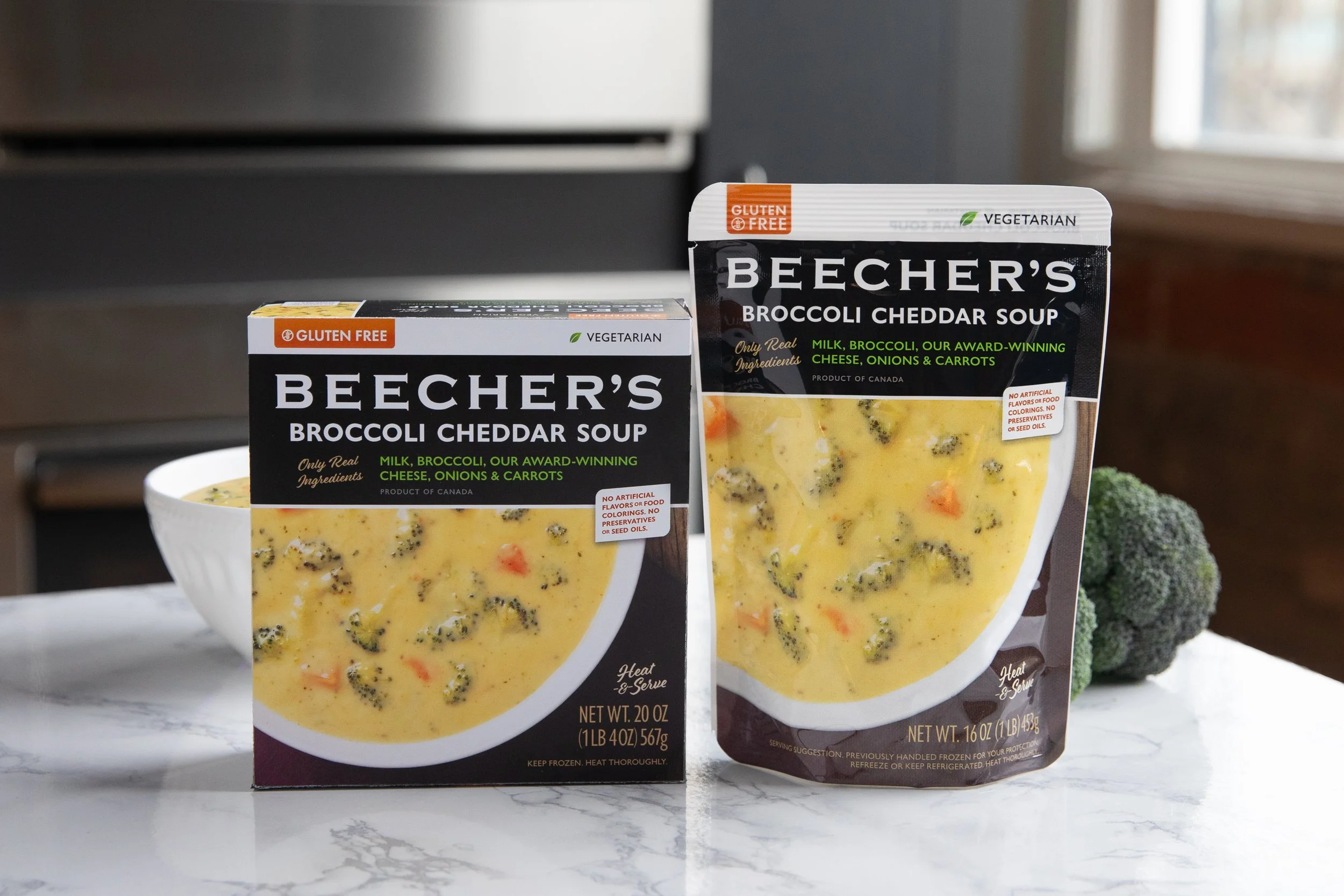 Two packages of Becher's Broccoli Cheddar Soup, one in a box and the other in a standing pouch, on a white marble surface with a broccoli floret in the background.