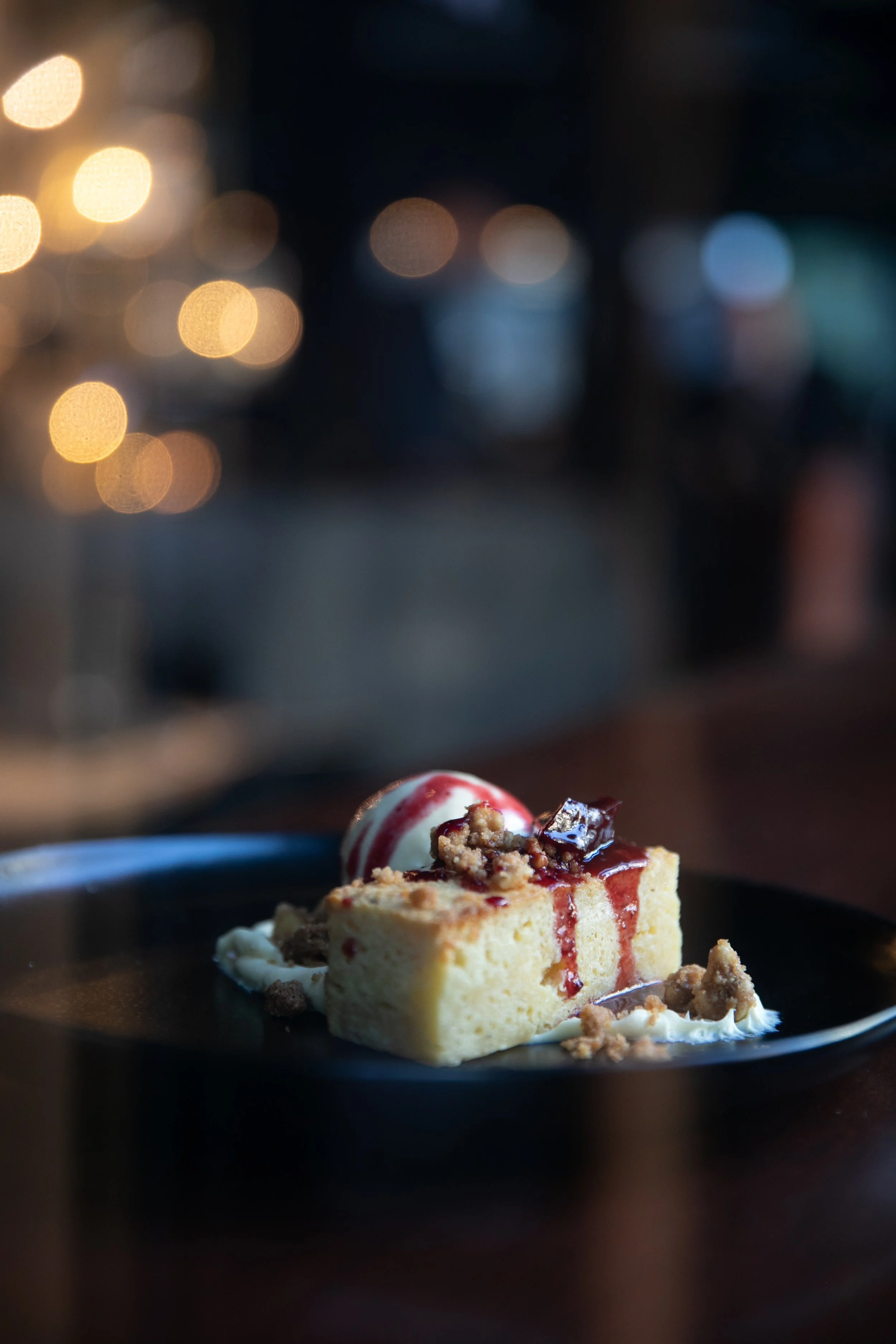 A plated dessert with a slice of cheesecake topped with red syrup, alongside a scoop of vanilla ice cream with red syrup, garnished with crumb pieces, on a black plate with a blurred background and warm bokeh lights.