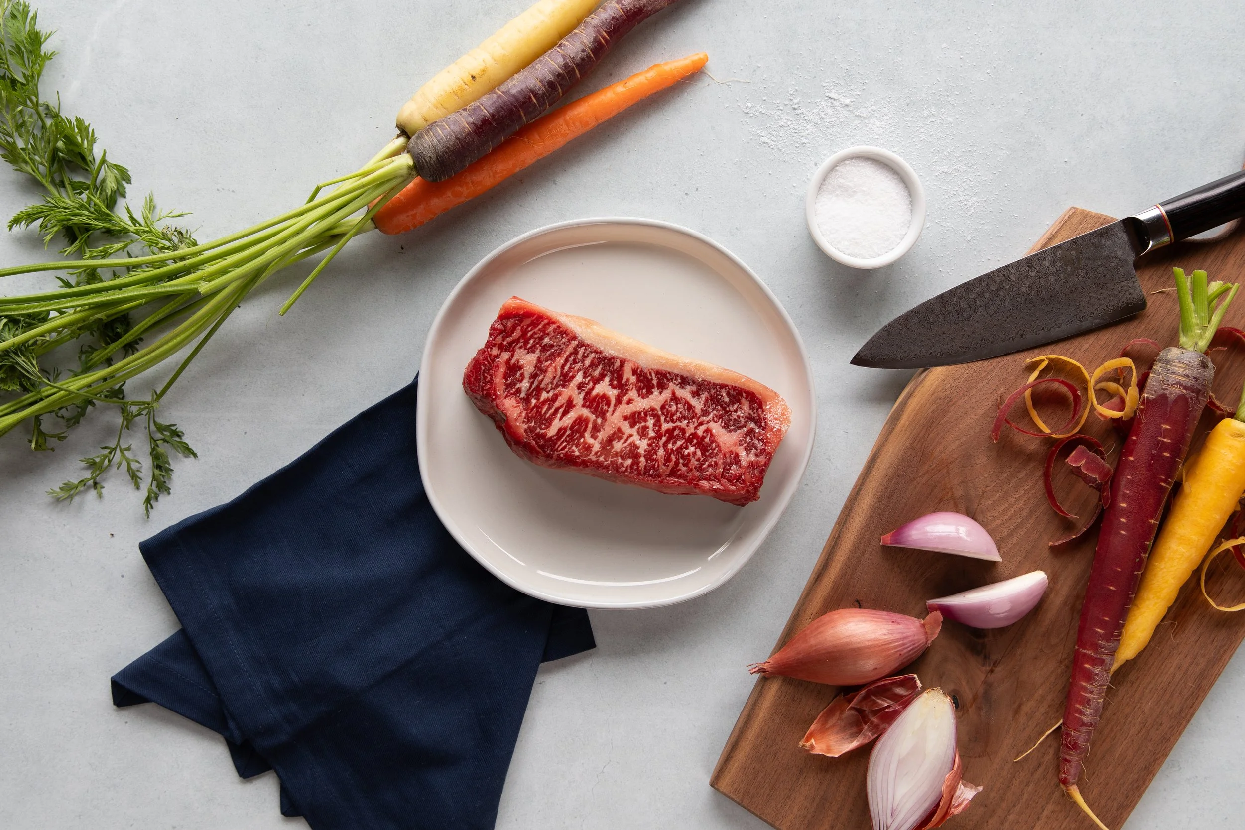 Raw marbled steak on a white plate surrounded by fresh vegetables including carrots, shallots, and garlic, with a cutting board, a chef's knife, and a small bowl of salt on a light gray surface.