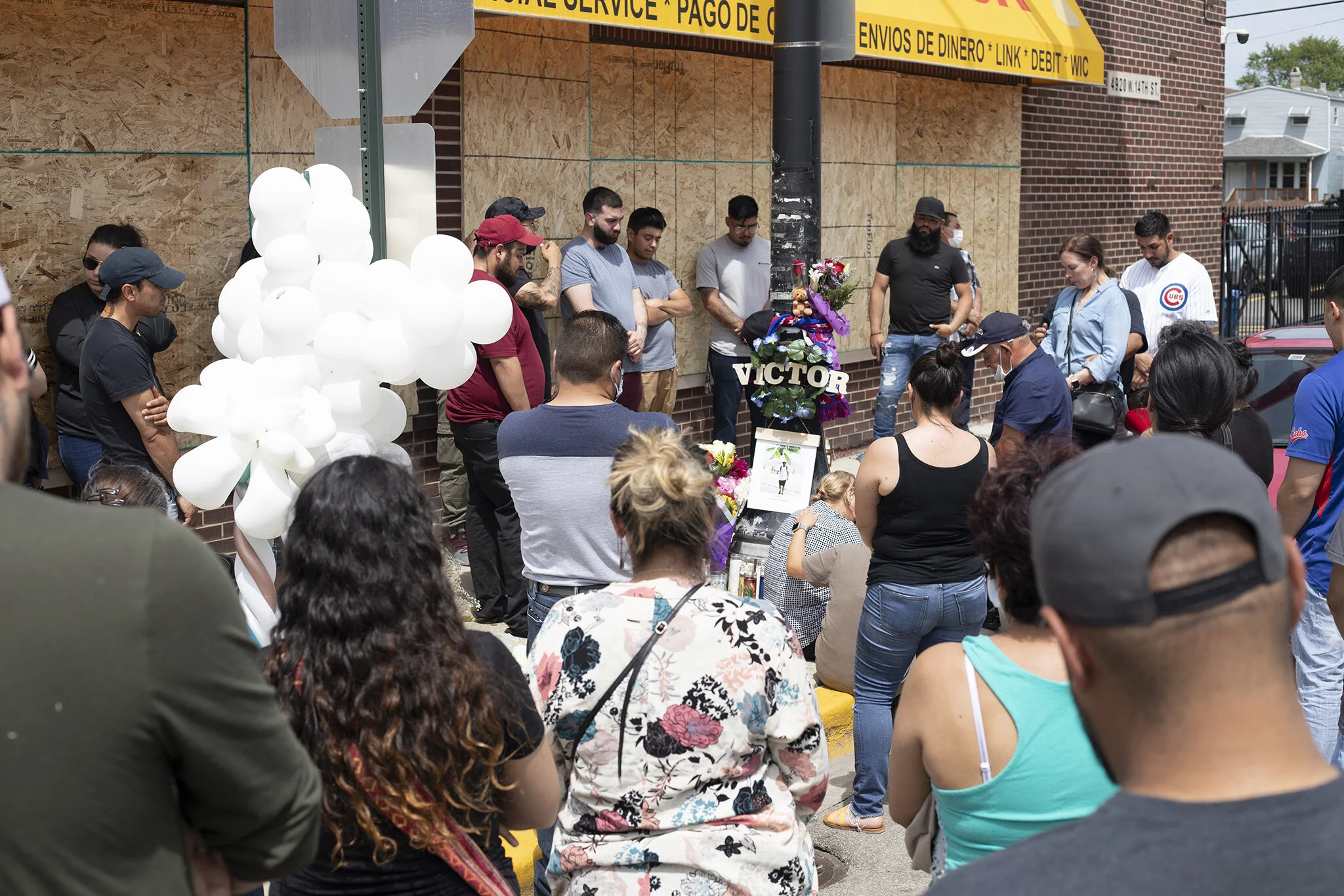 Image description: There is a crowd of people with their backs facing the camera. In the middle ground is a group of white balloons tied to a stop sign post on the left middle. In the center are flowers with a wooden sign of the name Victor attached to a black street post. Below that is a photo of the man who died from a gun shot wound and below on the cement are tall glass containers of candles. In the back ground is a grocery store with a yellow awning and boarded up windows and people facing the black street post with their heads bowed.