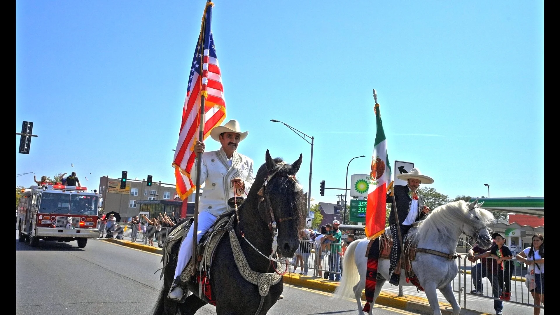 In a display of culture, two men ride on horseback with smiles and proud energy as one man holds an American flag and another holds a Mexican flag as they trot down the parade route with their horse.