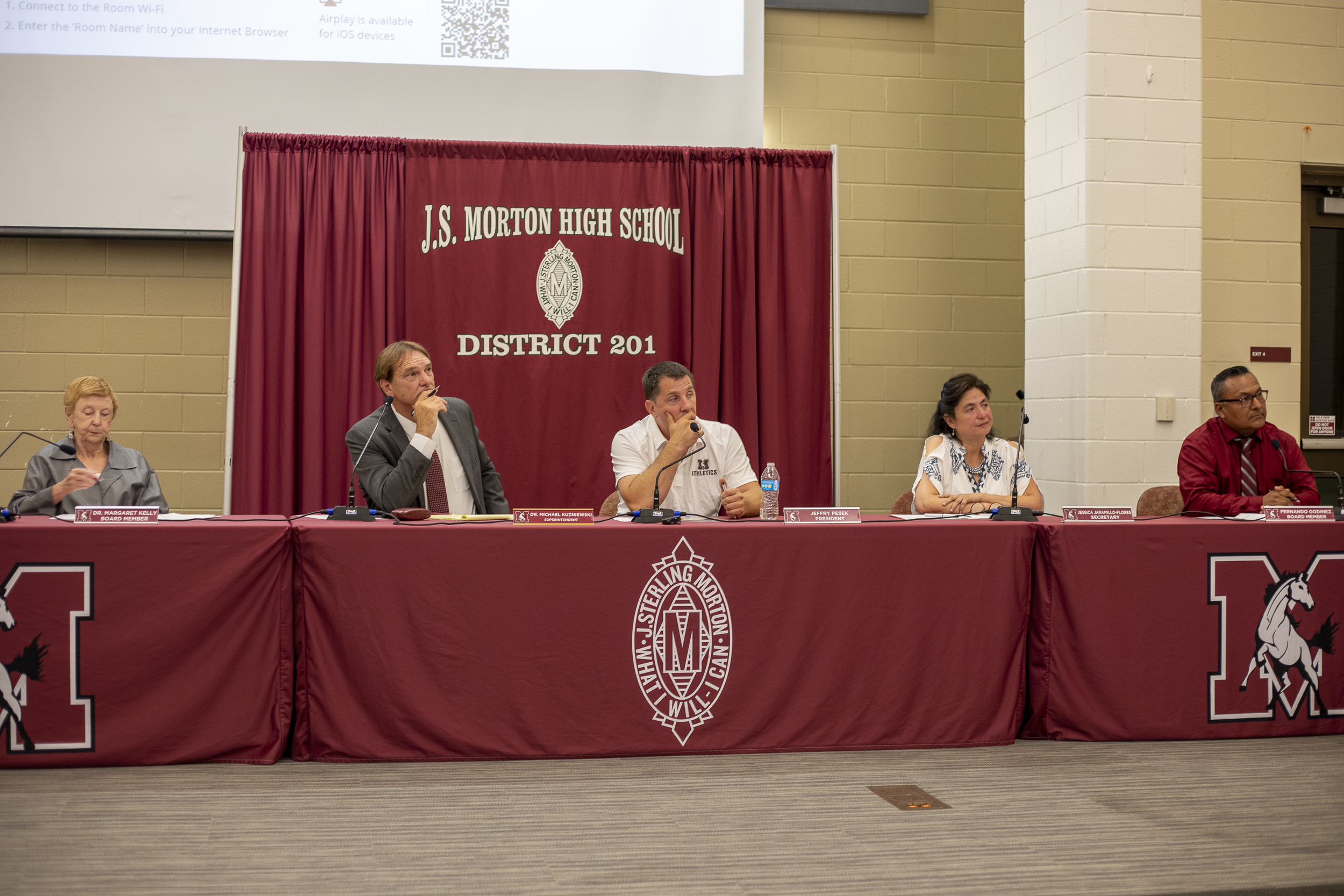 El consejo escolar del Distrito 201 se sienta a una mesa; el superintendente y el presidente del consejo se tapan la boca con la mano mientras observan a estudiantes, personal y miembros de la comunidad que dan sus comentarios.