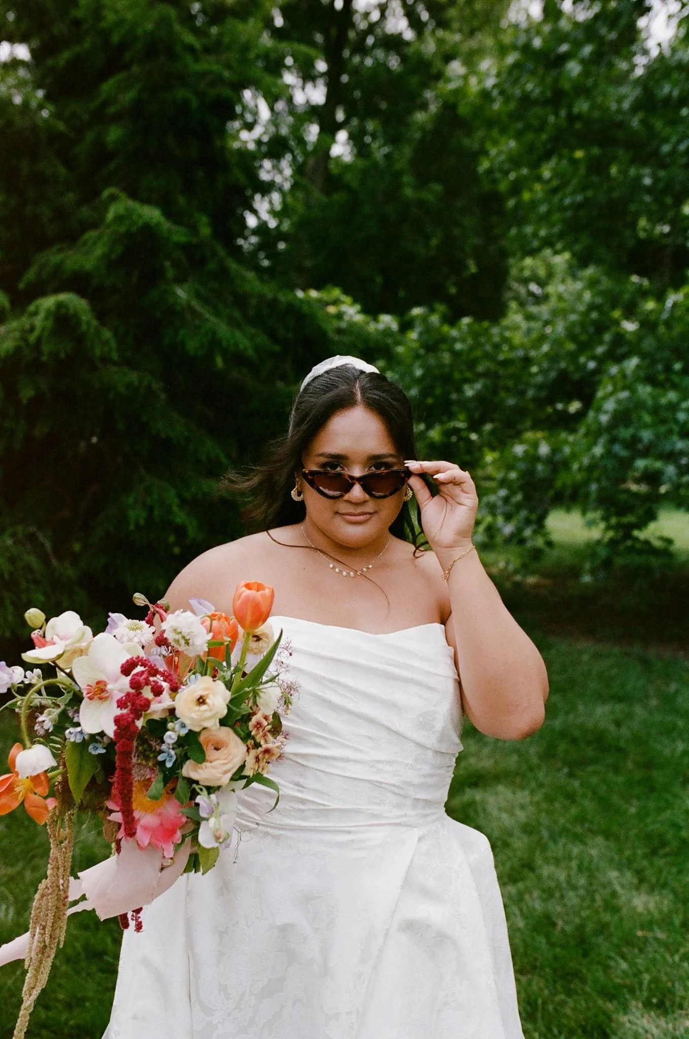 A woman in a white strapless dress holding a bouquet of flowers, wearing sunglasses and a headband, outdoors with green trees in the background.