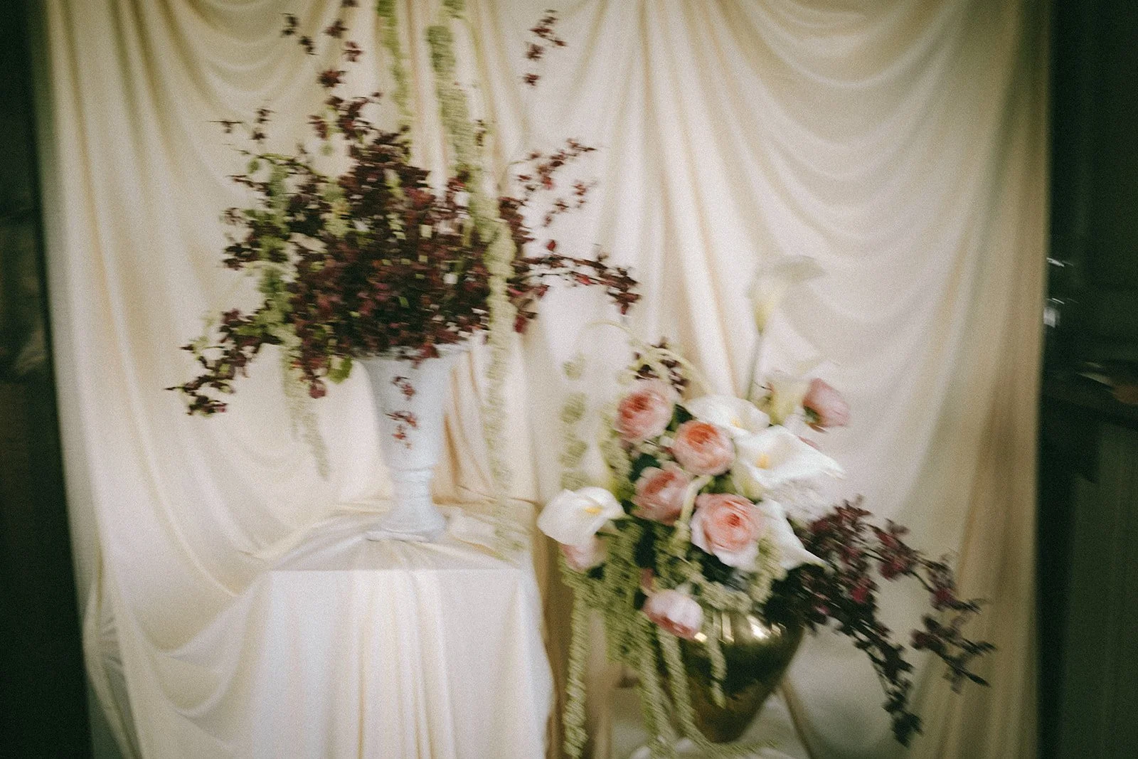 Elegant floral arrangement with pink roses, white calla lilies, and dark purple foliage in a black vase, placed on a cream-colored table with draped fabric and a matching backdrop.