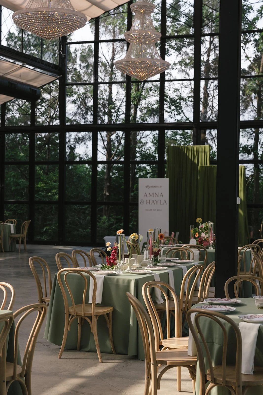Elegant indoor dining area with round tables covered with green tablecloths, decorated with floral arrangements, set with plates, glasses, and silverware. There are wooden chairs around the tables. Large windows reveal a lush green outdoor scenery. Two large chandeliers hang from the ceiling, and a sign with the names "Amna & Hayla" is visible in the background.