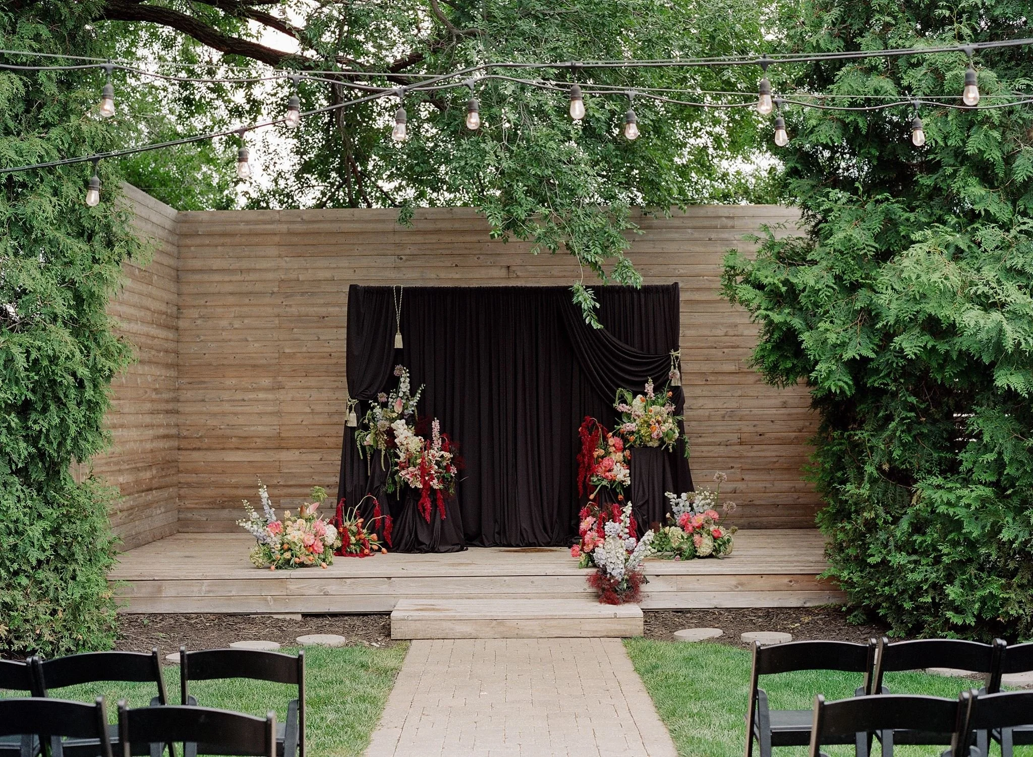 Outdoor wedding altar decorated with flowers, set against a wooden backdrop, with string lights hanging overhead, surrounded by green trees and chairs arranged for guests.