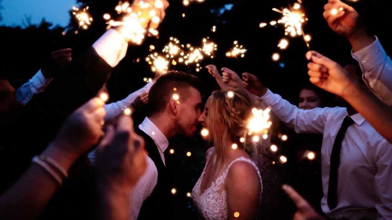 Newlywed couple sharing a romantic kiss surrounded by guests holding sparklers during a nighttime celebration at Barcaldine Castle