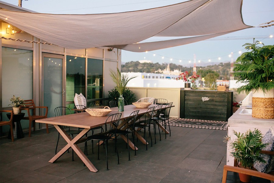 A beautiful rooftop terrace with a large wooden table, green plants, string lights, and sun sails to protect from the elements.